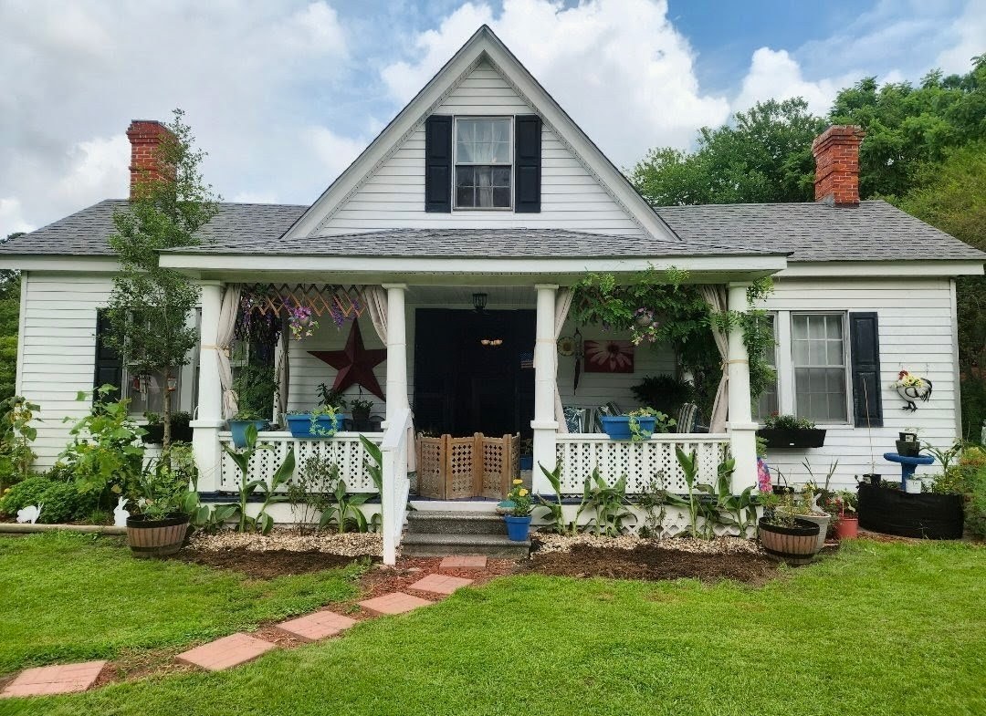 White farmhouse-style home with a front porch, dark shutters, and a large red star on the siding, set on a grassy lawn with trees framing the view belonging to Sponsored Residential Provider Angela Justice in Mathews, Virginia.