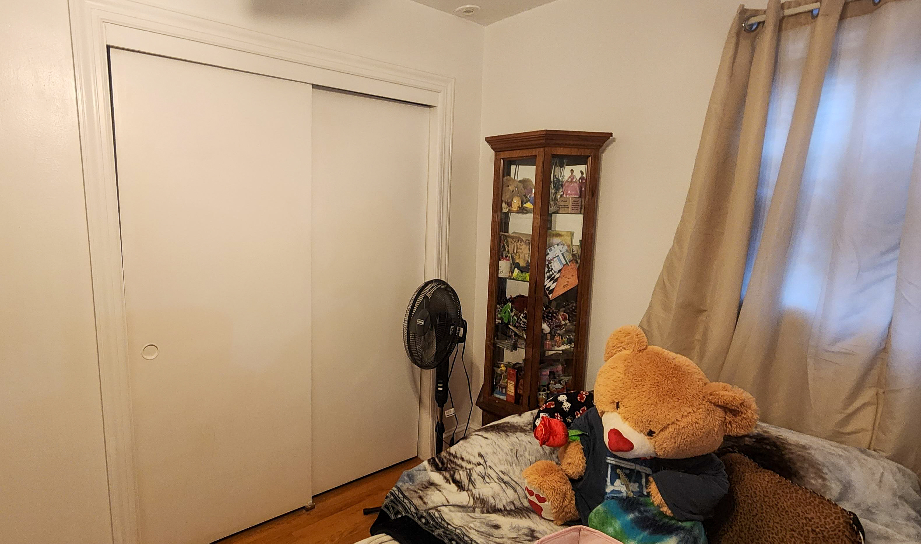 Bedroom corner with a sliding‑door closet, a tall glass display cabinet, and a sofa holding a large teddy bear near a window with beige curtains inside the home of Sponsored Residential Provider Cindy Freeman in Bassett, Virginia.
