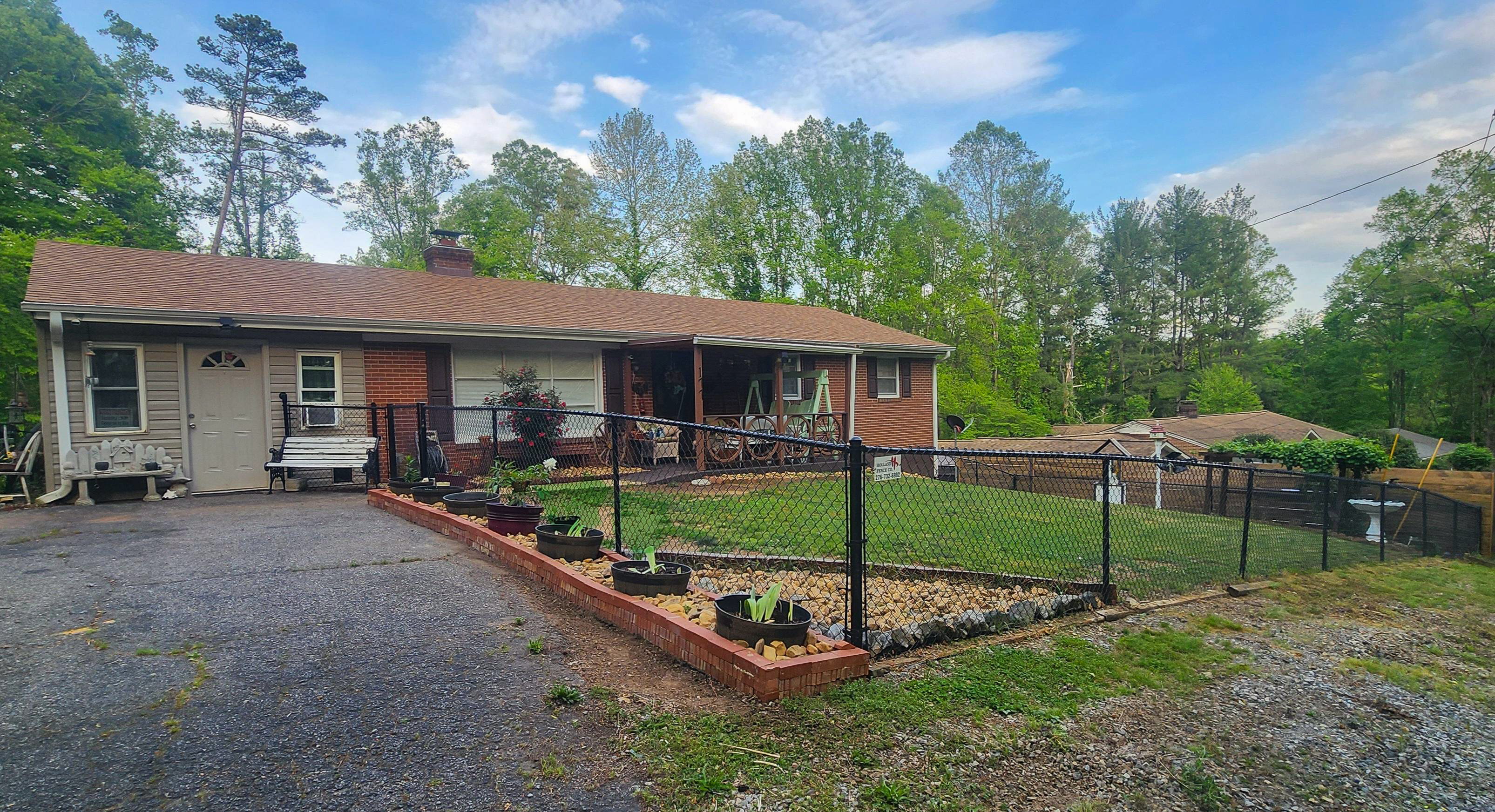Single‑story brick house with a covered porch, fenced garden area with plants, and a gravel driveway surrounded by trees  belonging to Sponsored Residential Provider Cindy Freeman in Bassett, Virginia.