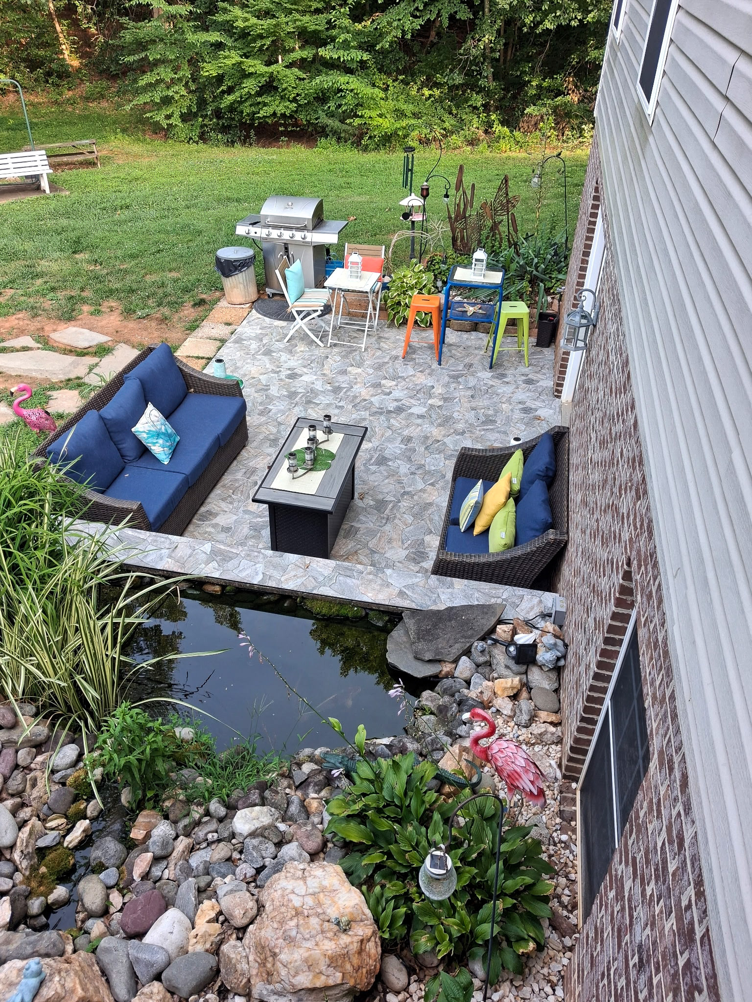 Stepping-stone patio with outdoor seating, grill, colorful stools, and a small pond bordered by rocks and plants in a backyard setting.