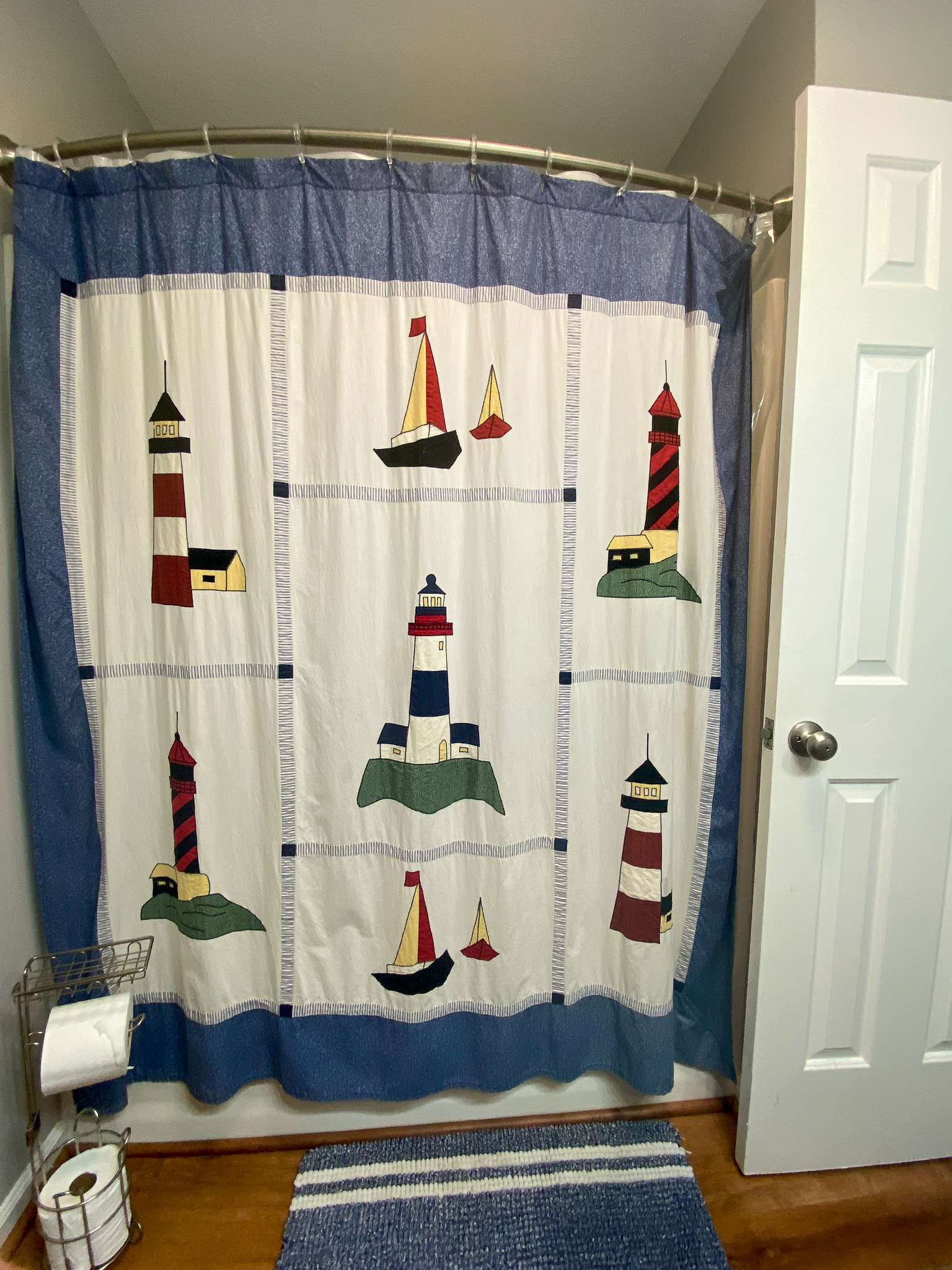 Shower curtain with colorful lighthouse and sailboat designs, blue border along the top and bottom, with wood‑look flooring and a white door to the right inside the home of Sponsored Residential Providers Karen and Rob Robertson in Verona, Virginia.