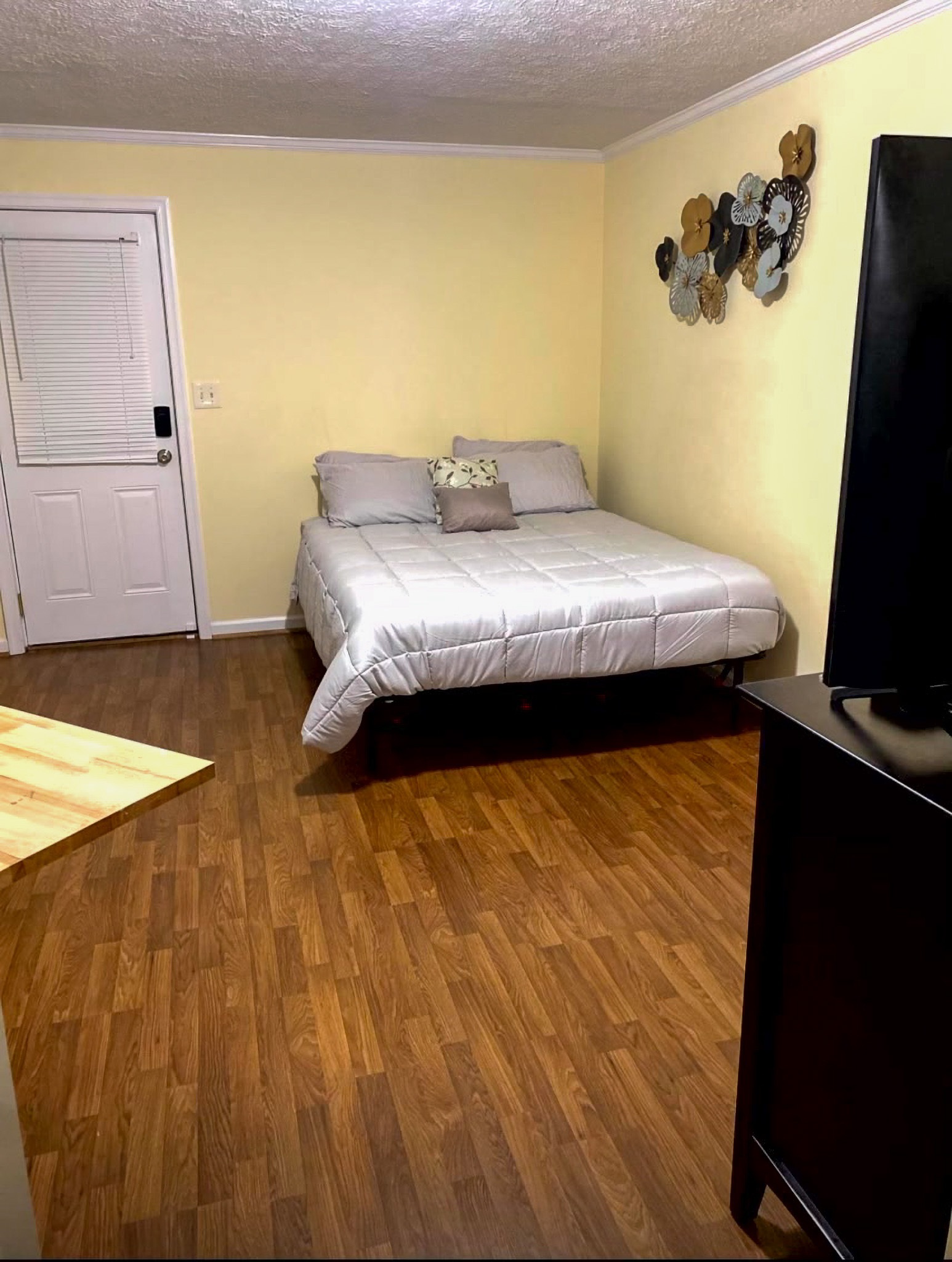 Bedroom with a low bed against a pale yellow wall, wood‑look flooring, wall décor above the bed, and a TV on a dark dresser in the foreground inside the home of Sponsored Residential Providers Takeyshia Gibson and Antonio Carter in Forest, Virginia.