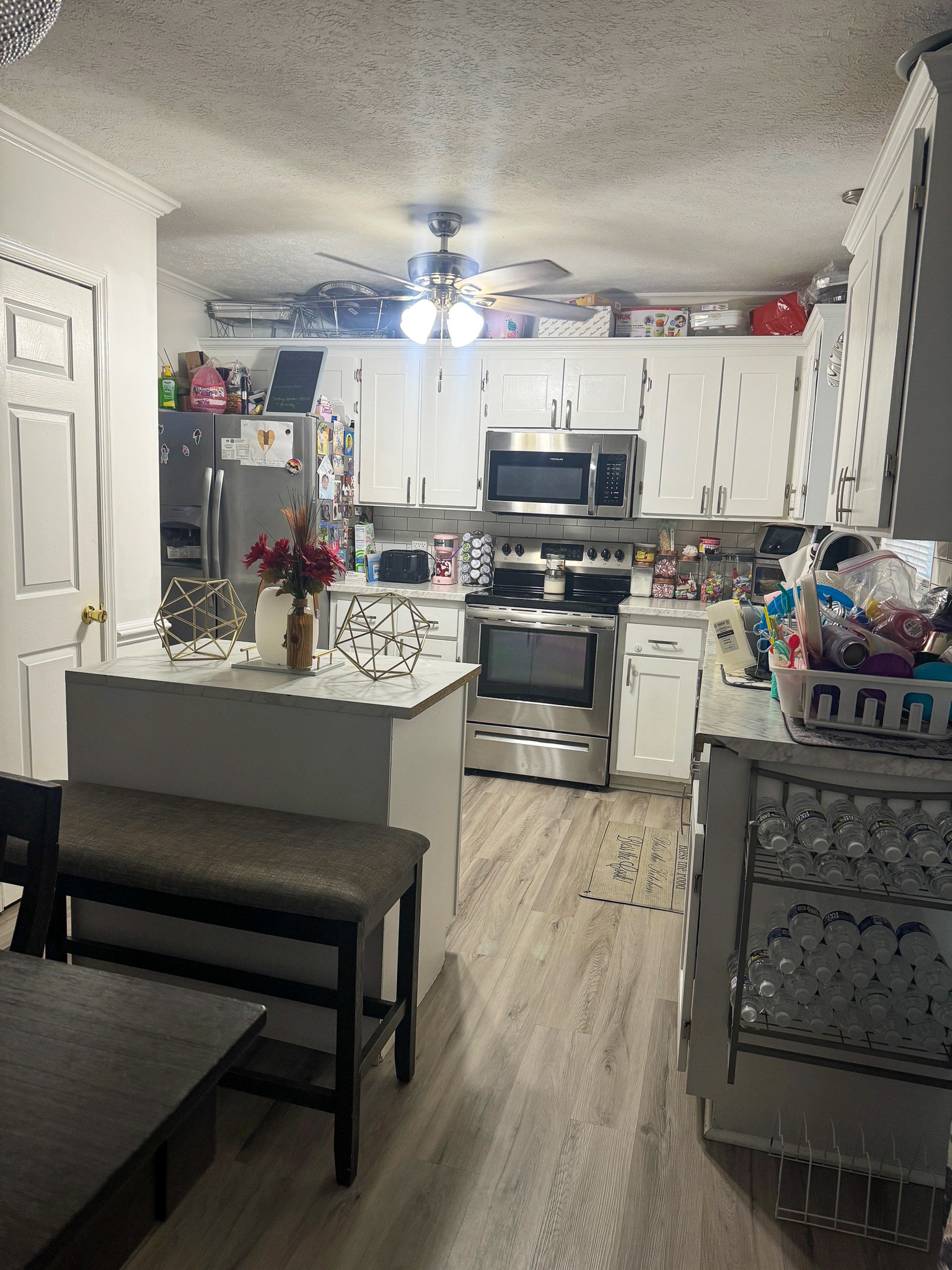 Compact kitchen with white cabinets, stainless‑steel appliances, a small island with flowers, and light wood flooring, with countertops holding various items inside the home of Sponsored Residential Providers Anthony Barnes and Demita Taylor in Forest, Virginia.