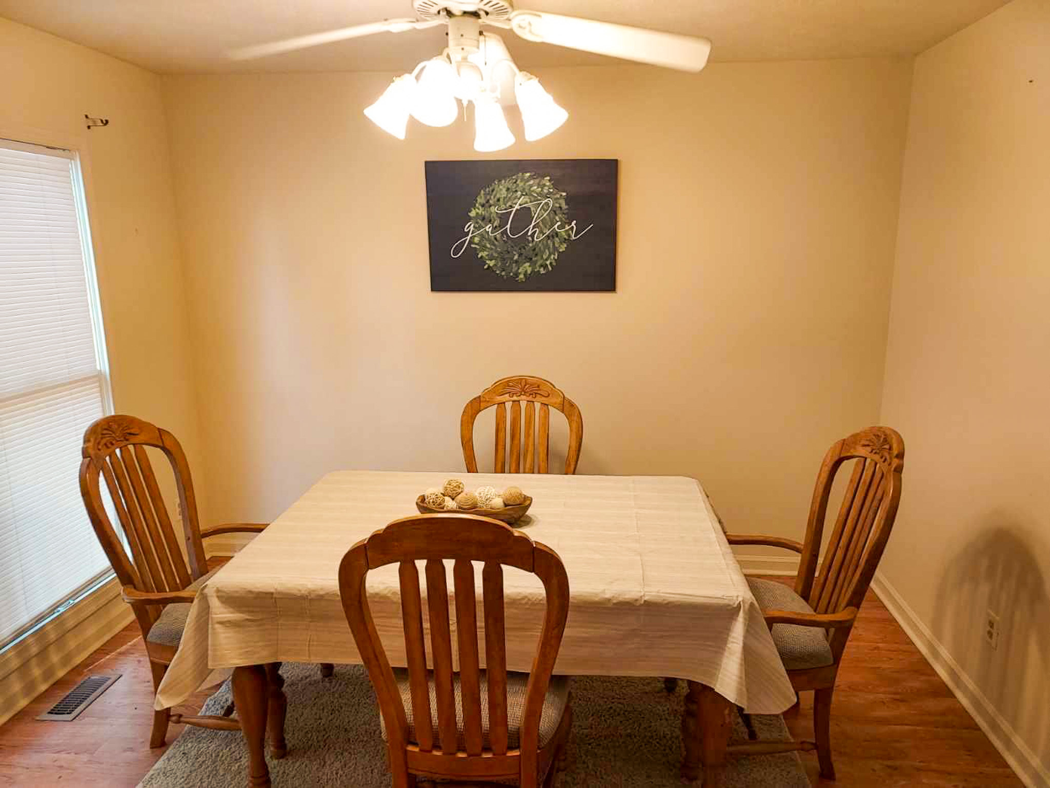 Dining room with a rectangular table covered in a white cloth, four wooden chairs, a ceiling fan with lights, and a wreath artwork on the wall inside the home of Sponsored Residential Provider David Wilcox in Harrisonburg, Virginia.
