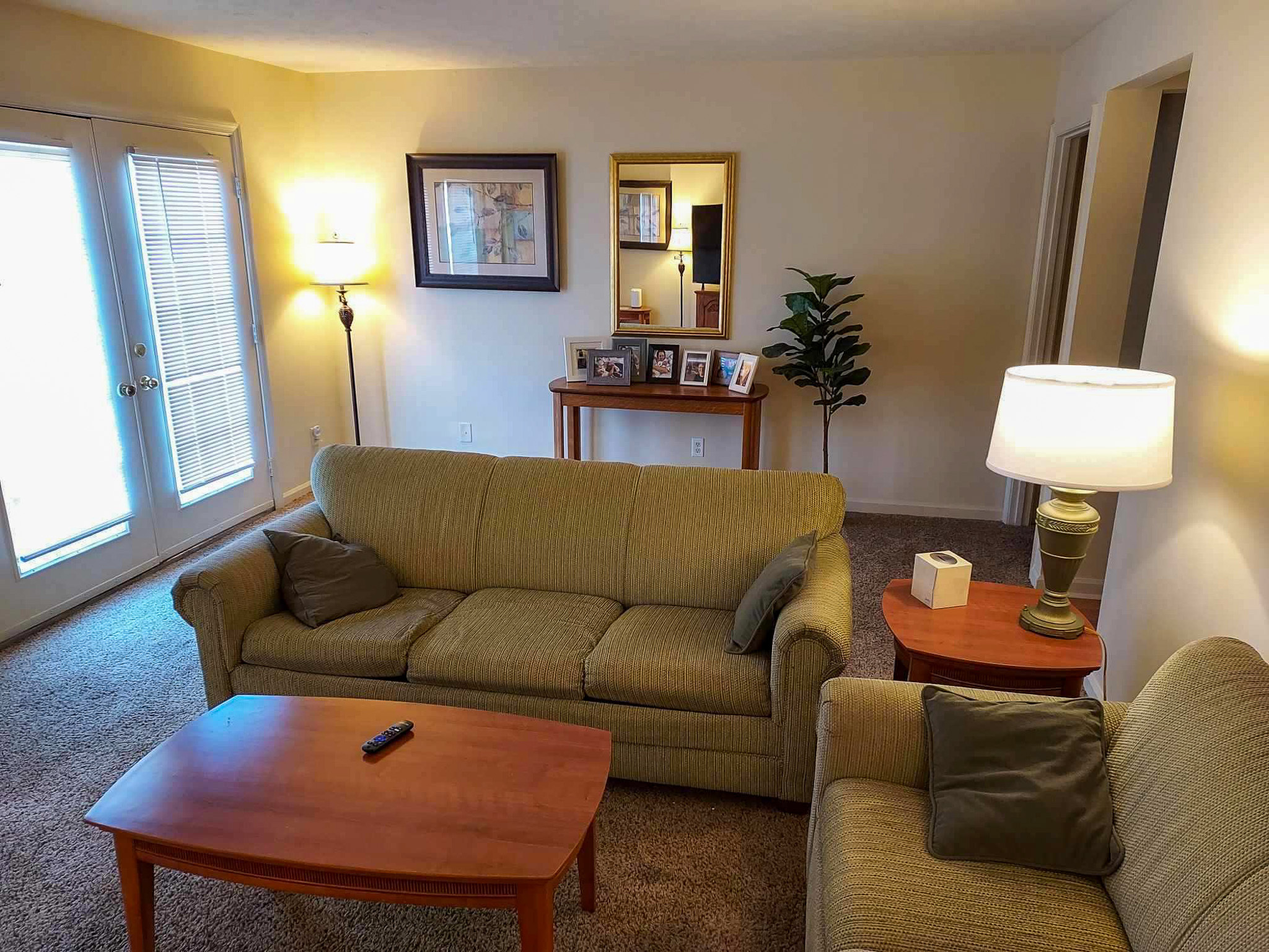 Living room with two green sofas, a wooden coffee table, end tables with lamps, and a console table with framed photos beneath a wall mirror inside the home of Sponsored Residential Provider David Wilcox in Harrisonburg, Virginia.