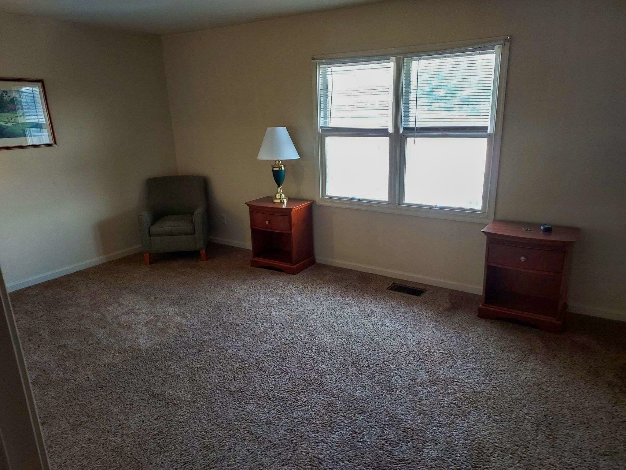 Carpeted room with double window, a wooden side table with a lamp, an empty wooden nightstand, and a single armchair in the corner inside the home of Sponsored Residential Provider David Wilcox in Harrisonburg, Virginia.
