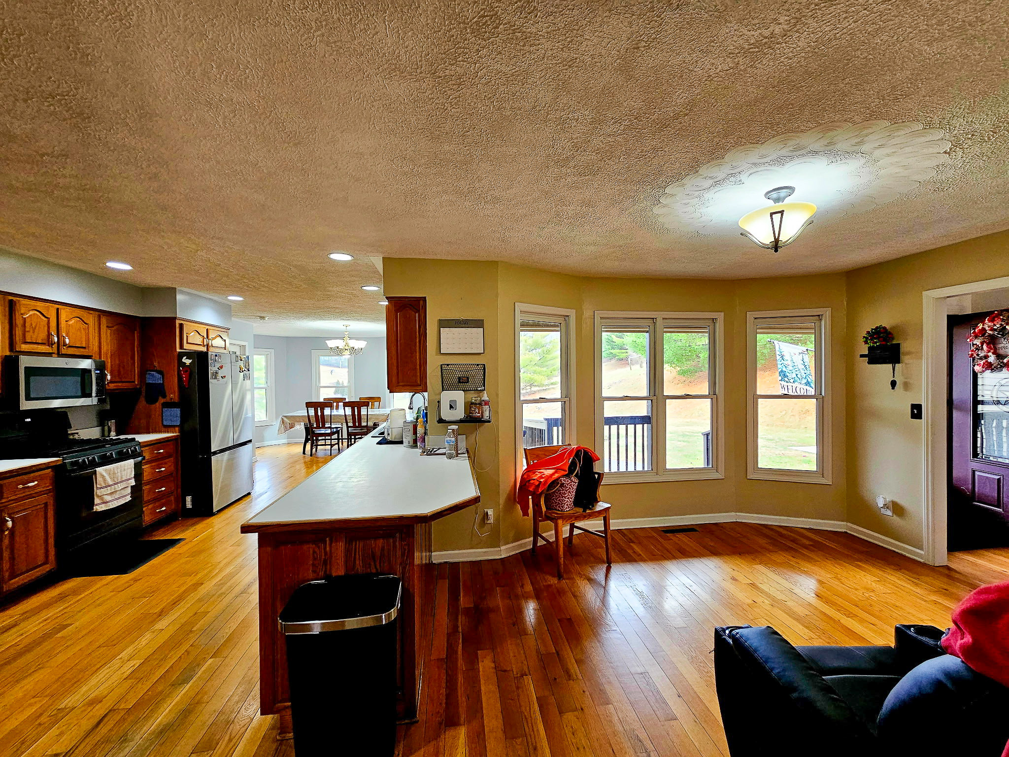 An open kitchen and dining area with wood floors, dark wood cabinets, a large white peninsula counter, and a row of windows overlooking the yard inside the home of Group Home Provider Dylan Mays in Woodlawn, Virginia.