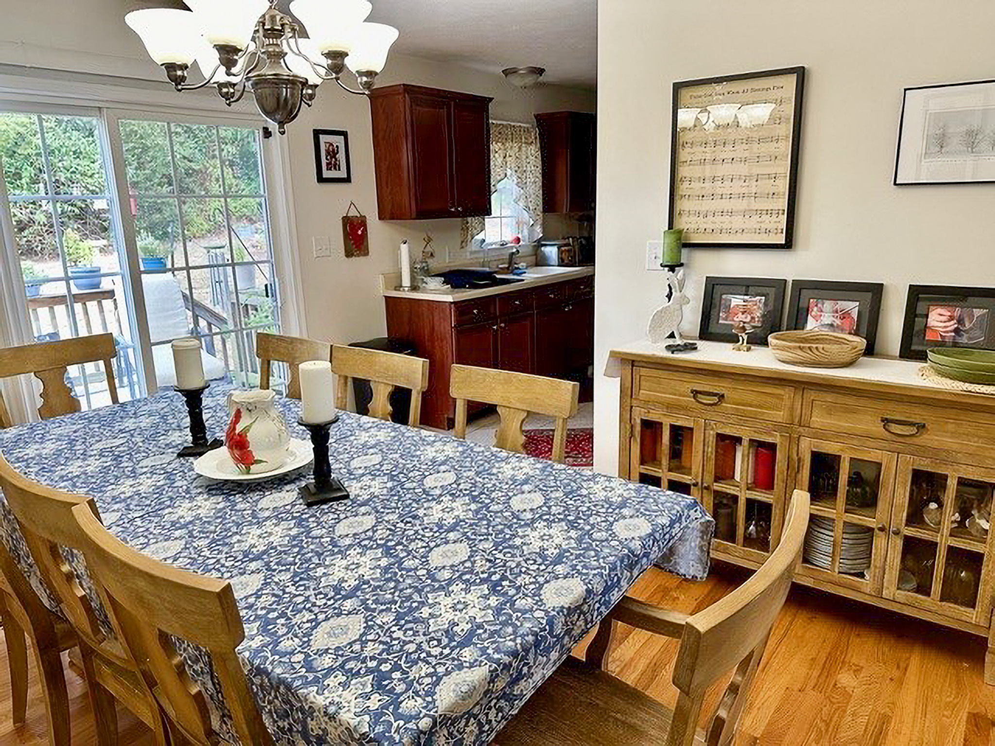 Dining room with a blue patterned tablecloth, wooden chairs, sideboard with framed decor, and a view into a kitchen with dark wood cabinets inside the home of Sponsored Residential Providers Nancy and Zach Kaplan in Waynesboro, Virginia.