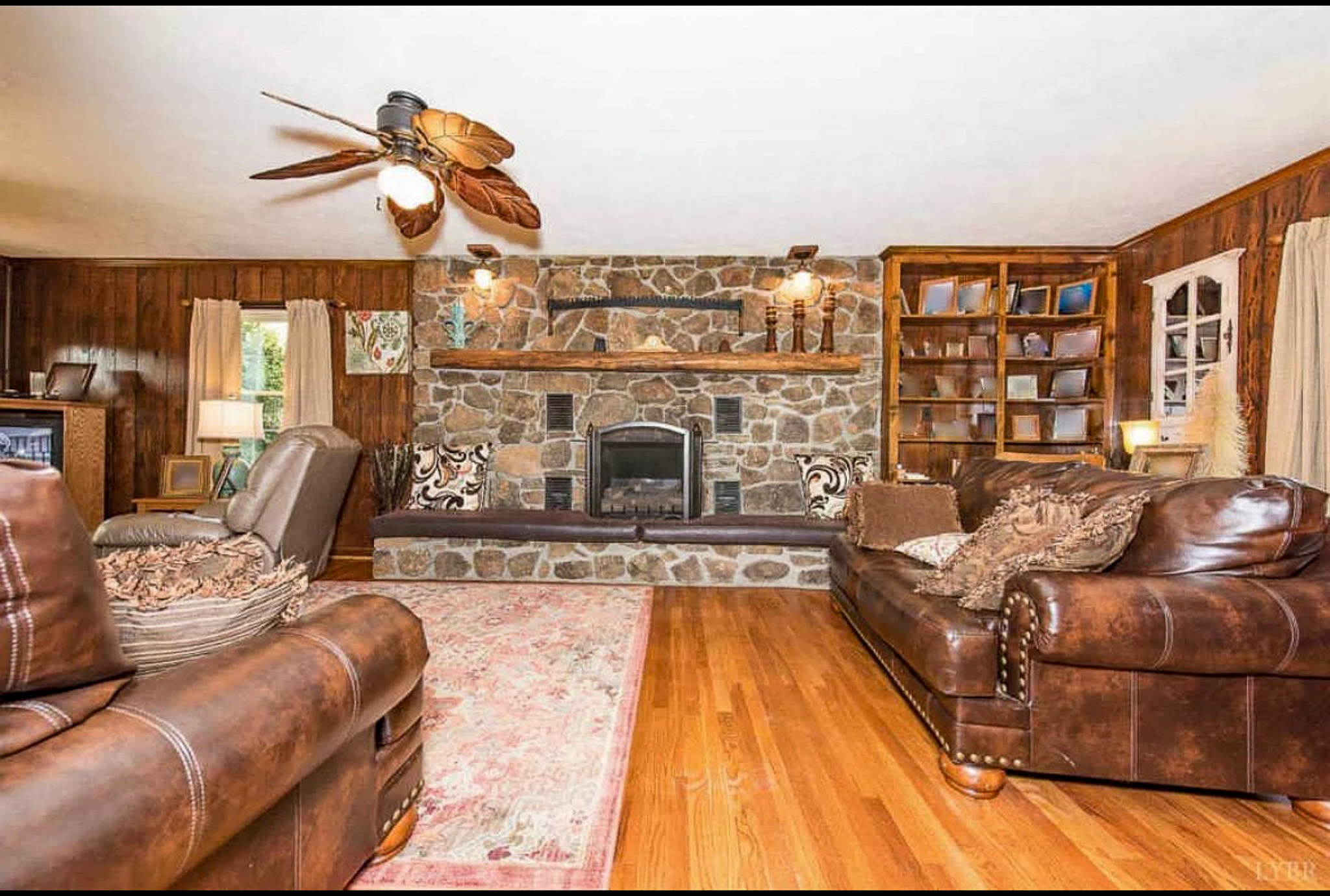 A warm living room with wood paneling, leather sofas, a large stone fireplace with a mantel, built‑in shelves, and hardwood floors inside the home of Sponsored Residential Providers Carolyn and Phil Ware in Amherst, Virginia.