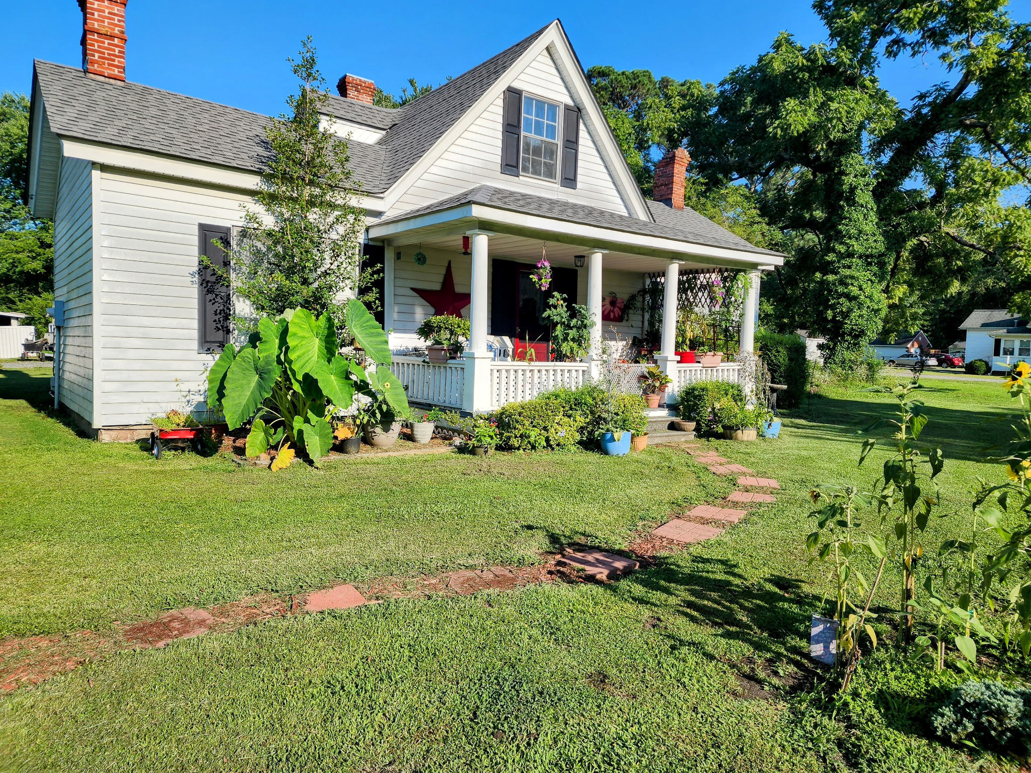 White farmhouse with a covered front porch, potted plants, and a landscaped yard under a bright blue sky belonging Sponsored Residential Provider Angela Justice in Mathews, Virginia.