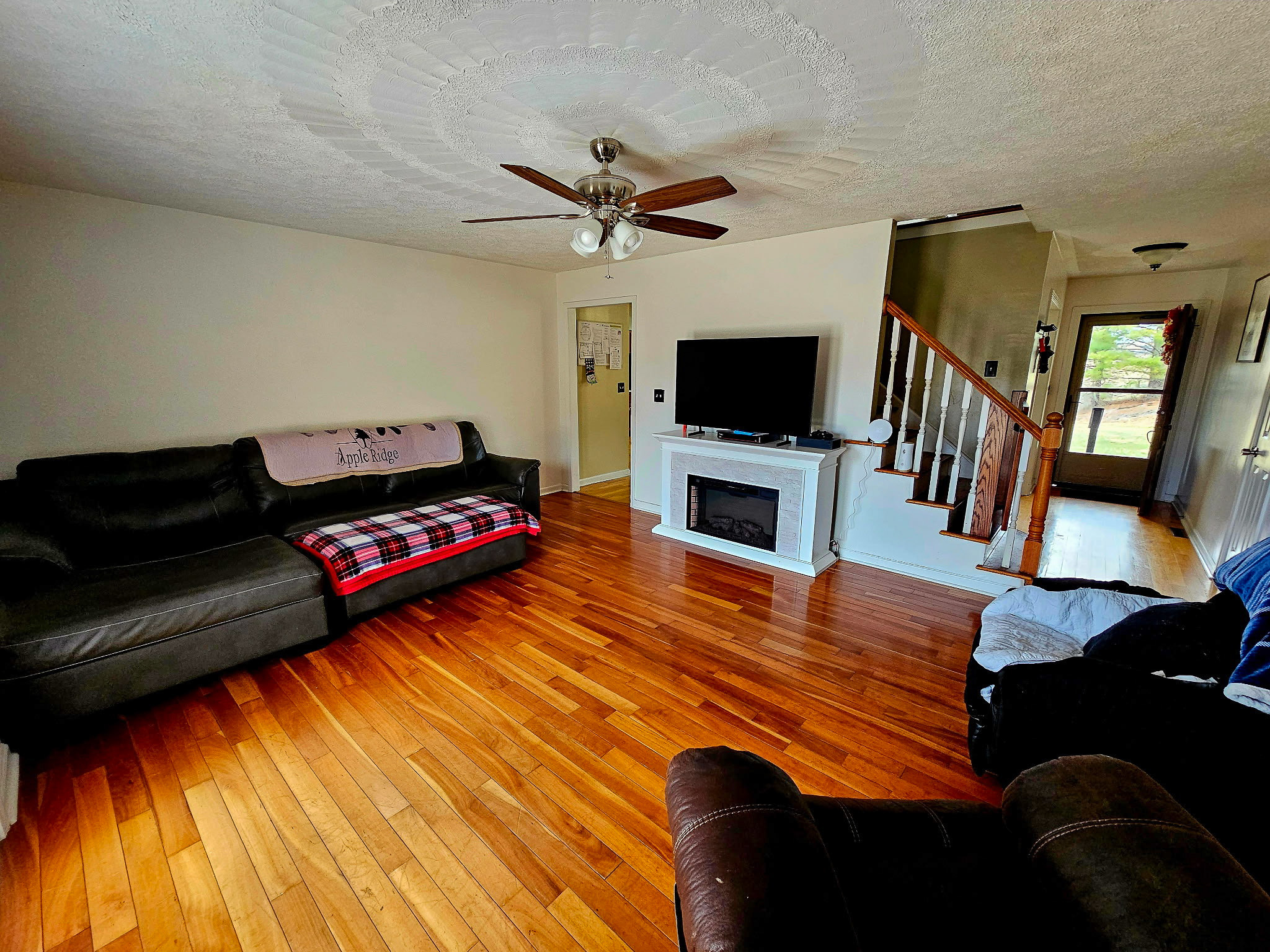 A bright living room with wood floors, dark sectional sofas, a ceiling fan, and a TV on a white fireplace-style console near a staircase and front entryway inside the home of Group Home Provider Dylan Mays in Woodlawn, Virginia.