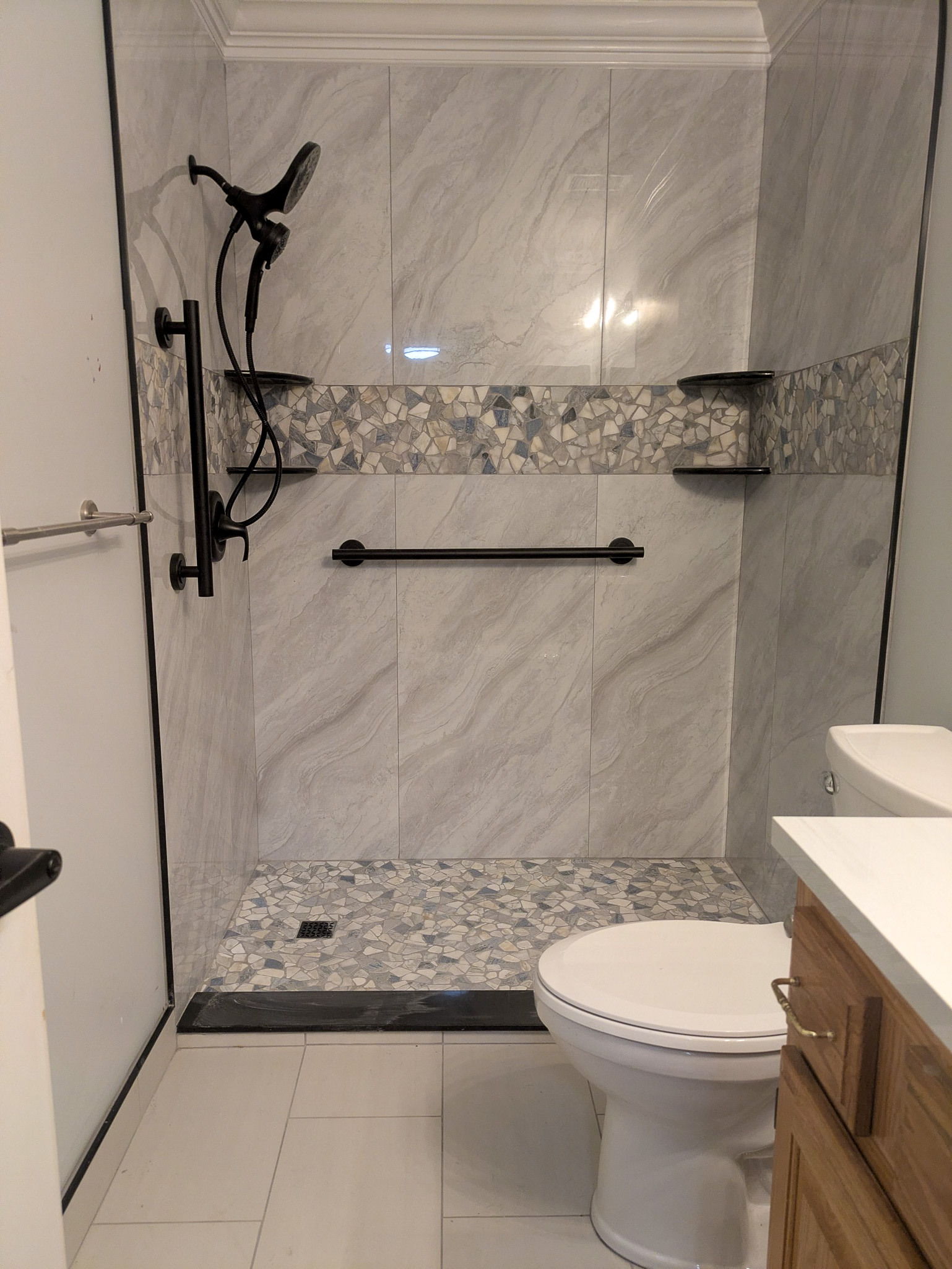 Shower enclosure with marble-style wall tiles, mosaic horizontal accent strip, matte black grab bar, corner shelves, and a handheld shower head.