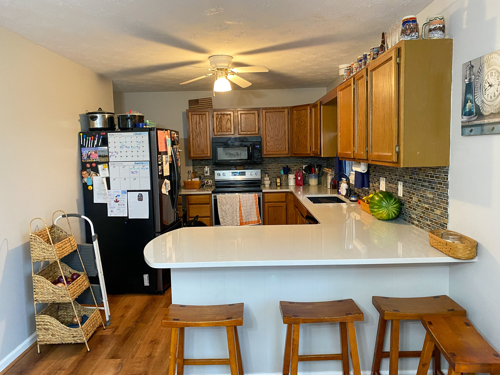 Kitchen with wood cabinets, white countertops, tile backsplash, stainless steel appliances, black refrigerator with notes and magnets, and a breakfast bar with three wooden stools inside the home of Sponsored Residential Providers Karen and Rob Robertson in Verona, Virginia.