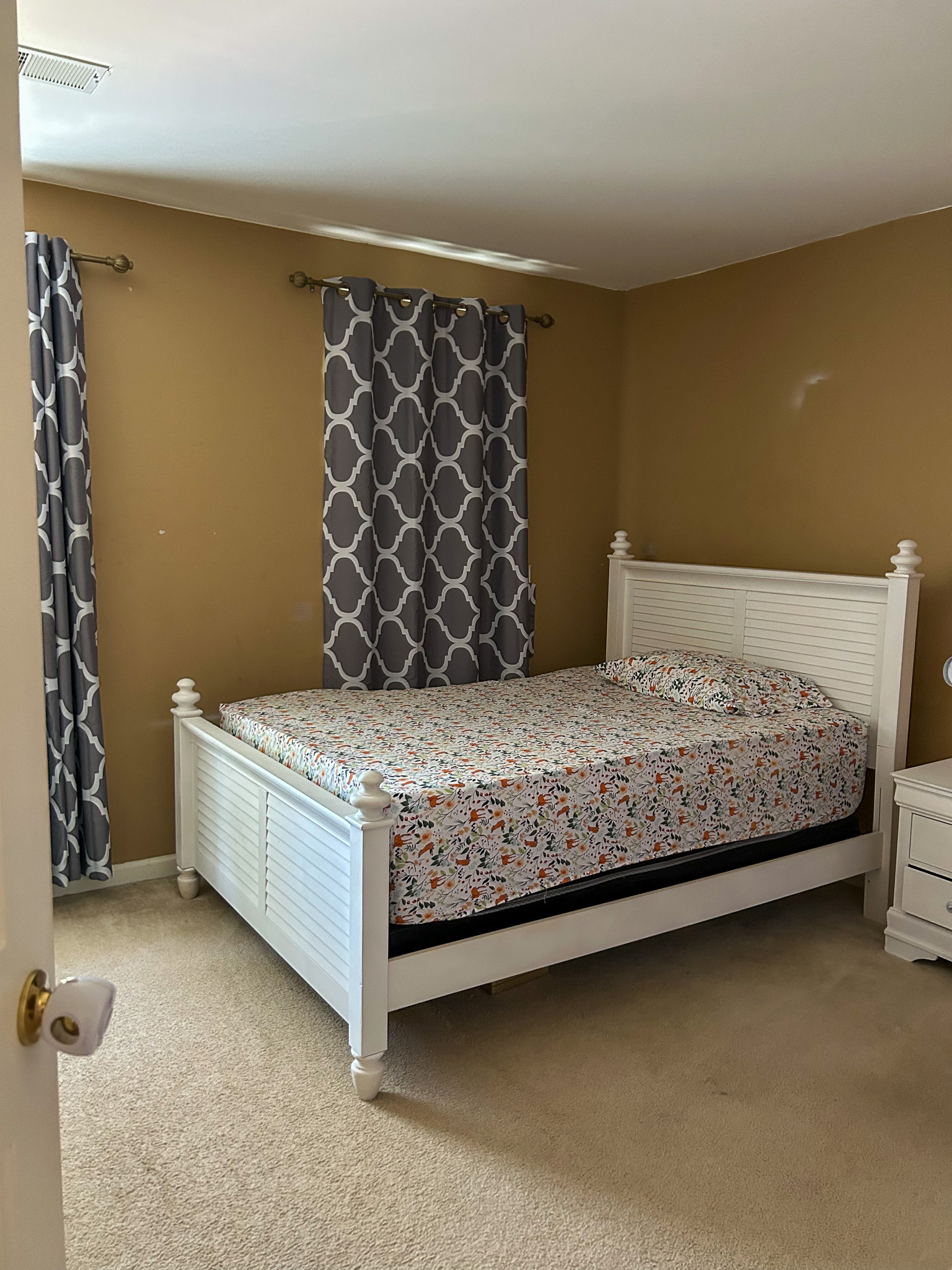 Bedroom with white bed frame, patterned bedding, carpeted floor, and curtain-covered windows inside the home of Sponsored Residential Provider Aidah Brassier in Spotsylvania, VA.