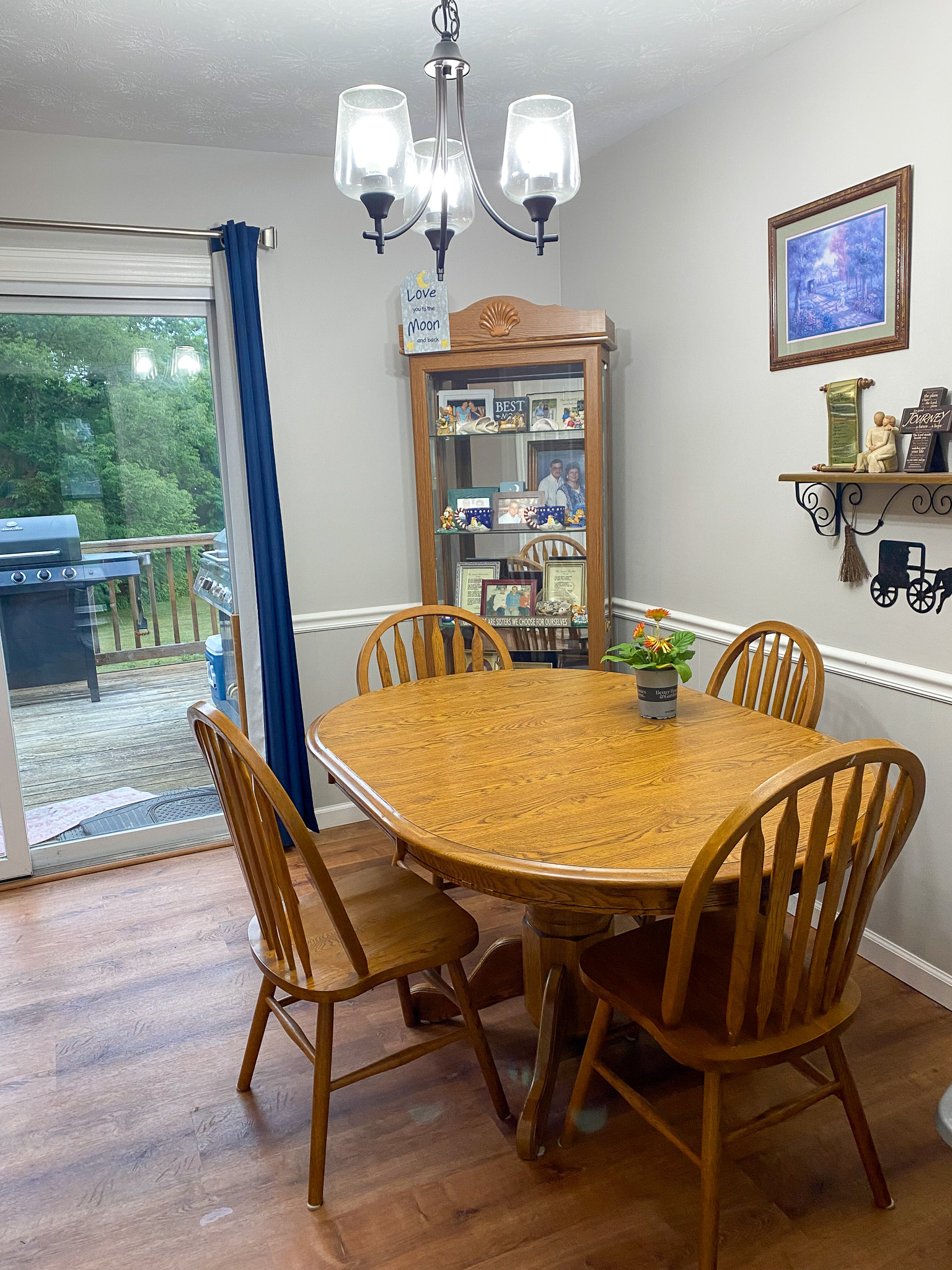 Dining area with a wooden table and four matching chairs, chandelier overhead, sliding glass door leading to a deck, wall shelf with decor, and a glass-front cabinet inside the home of Sponsored Residential Providers Karen and Rob Robertson in Verona, Virginia.