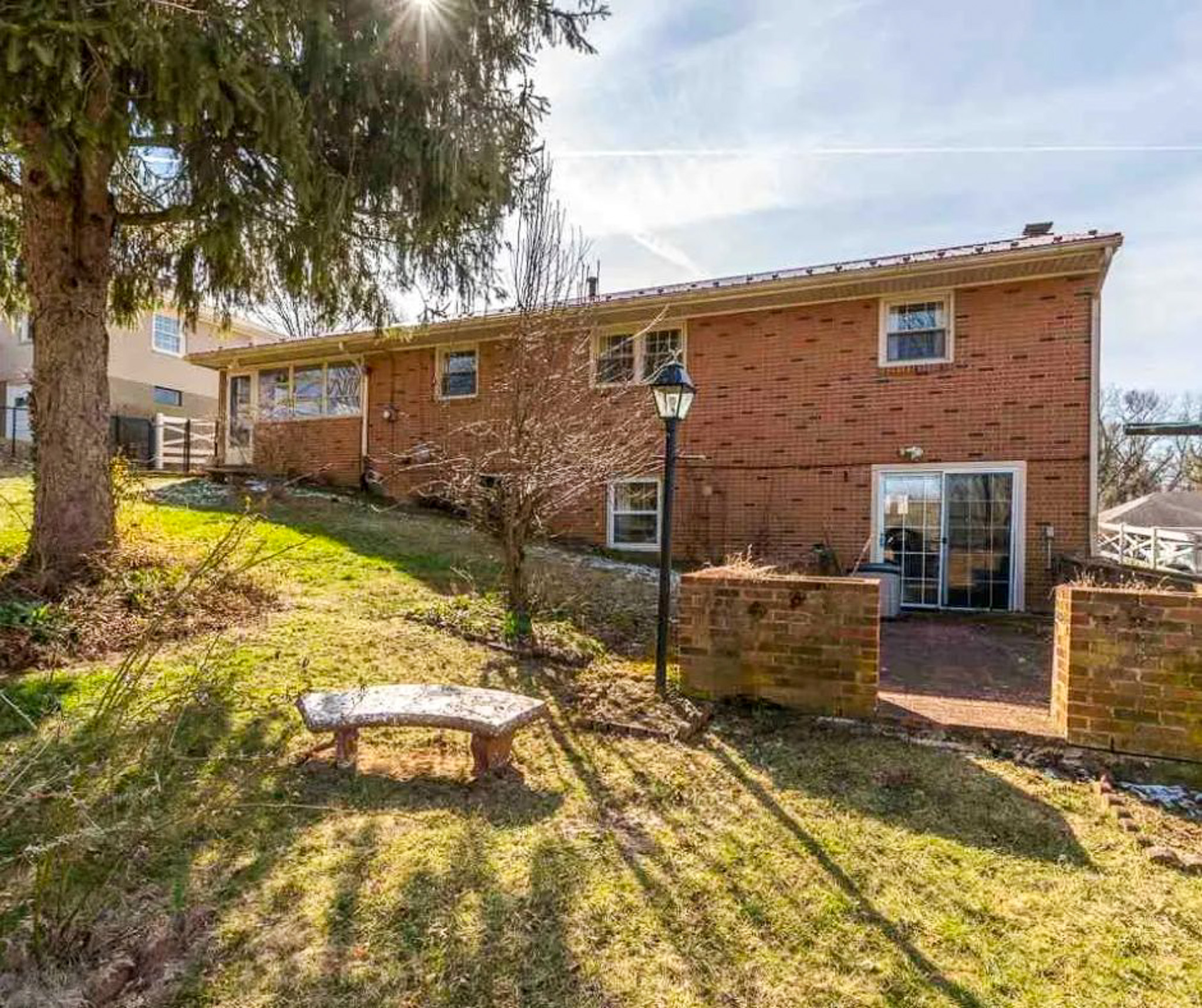 Brick home with a walk‑out lower level, sliding glass door, small patio, and sloped grassy yard with a large tree, lamp post, and a bench in the foreground at the home of Sponsored Residential Providers Bill and Heather Terry in Staunton, Virginia.