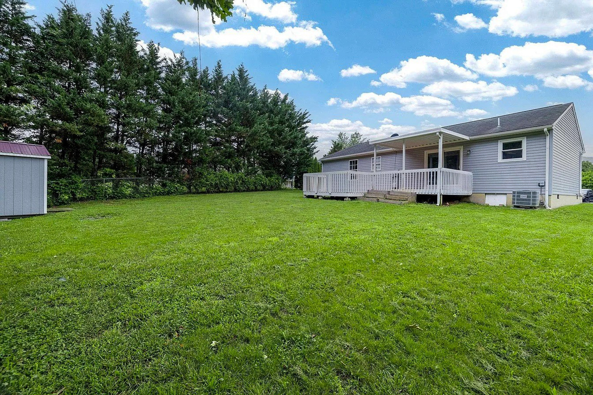 Back view of a gray single‑story house with a covered porch, large grassy yard, tall evergreen trees along the property line, and a small shed to the left  belonging to Sponsored Residential Provider Gabrielle Straway in Grottoes, Virginia.