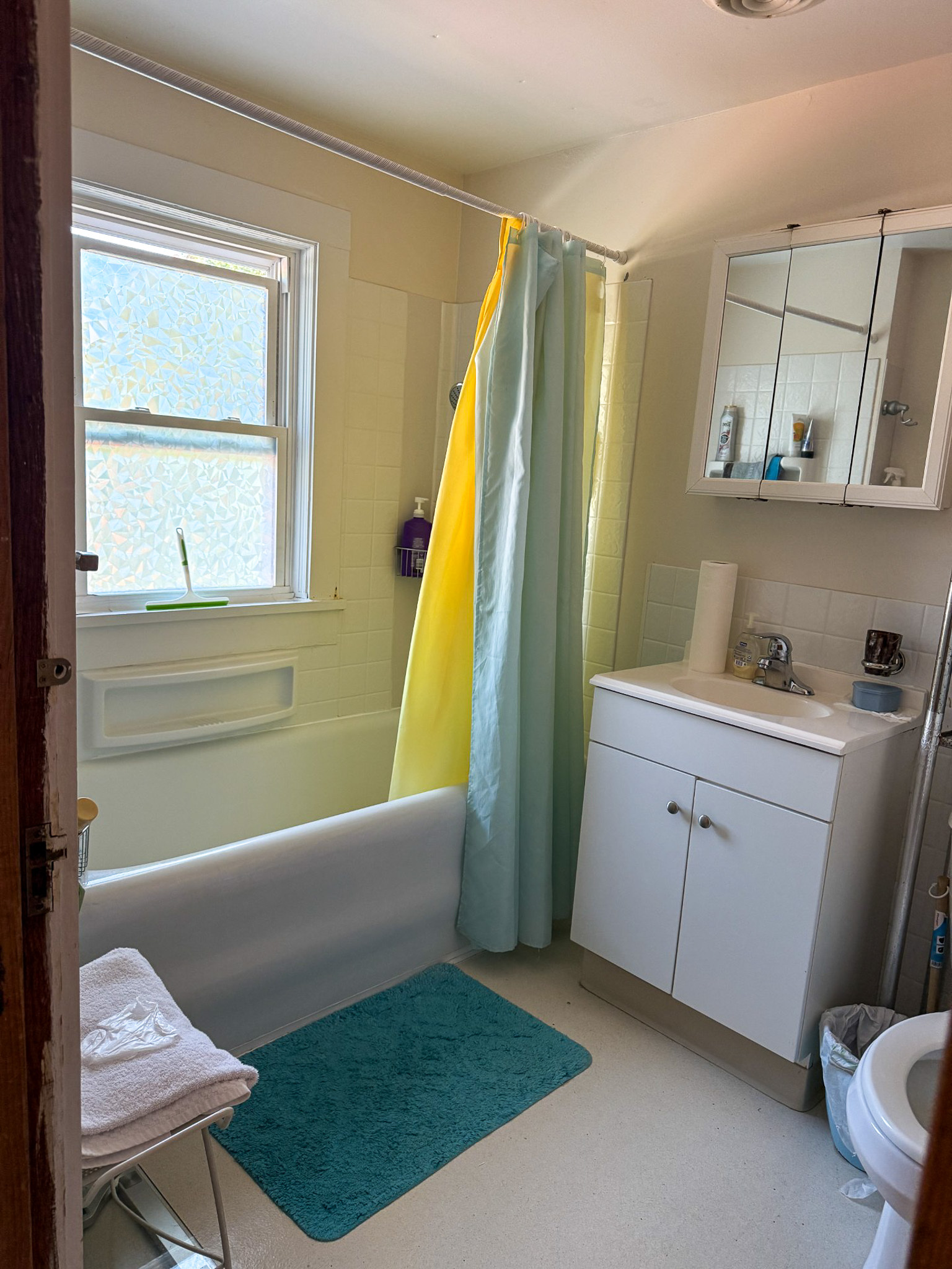 Small bathroom with a bathtub and colorful shower curtain, white vanity with mirror cabinet, window over the tub, and teal bath mat inside the home of Sponsored Residential Provider Tammy Hunt in Floyd, Virginia.