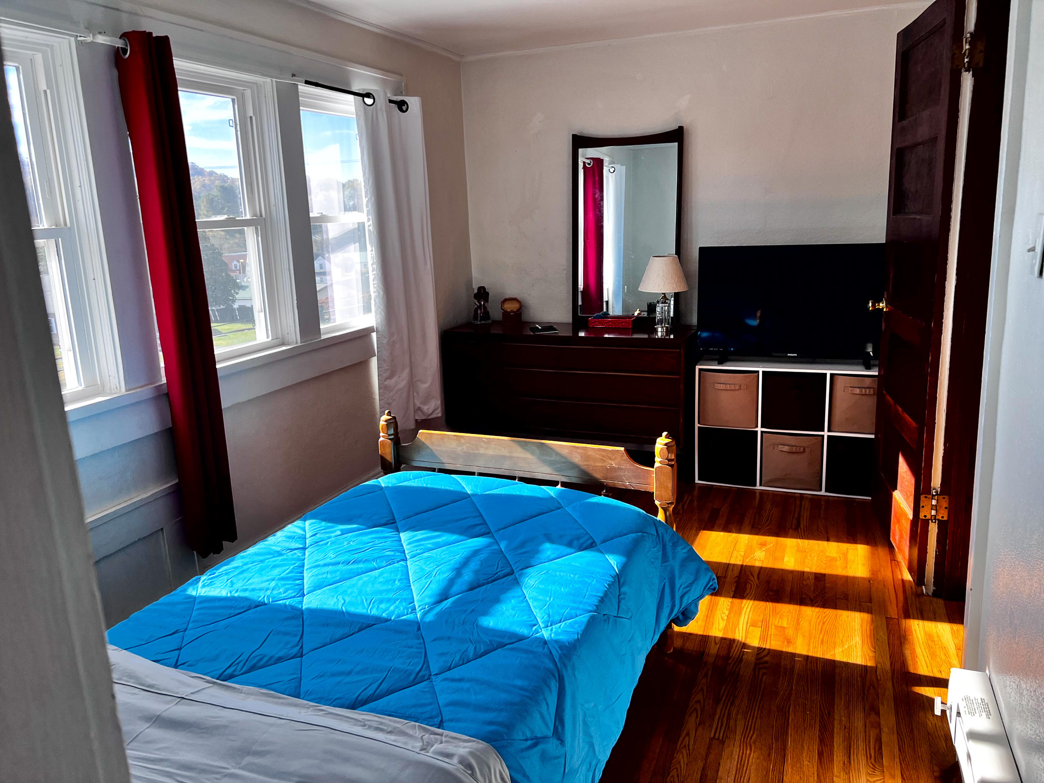 Bright bedroom with a wooden bed frame, blue comforter, dresser with mirror, cube storage, and multiple windows letting in sunlight inside the home of Sponsored Residential Provider Tammy Hunt in Floyd, Virginia.