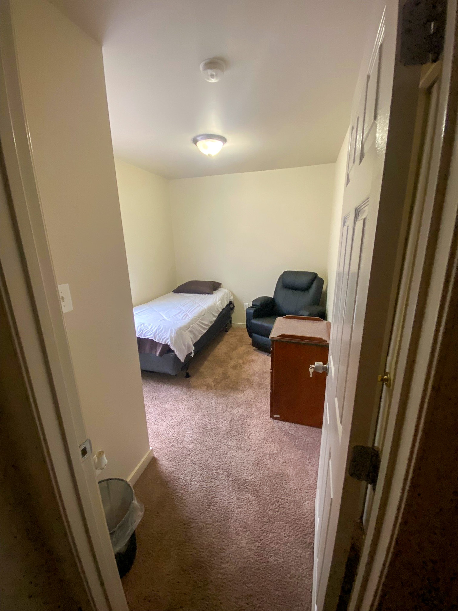 A simple bedroom with a bed, dresser, and padded chair positioned around the walls inside the home of Sponsored Residential Providers Xaiver Heath and David Williams in Richmond, Virginia.