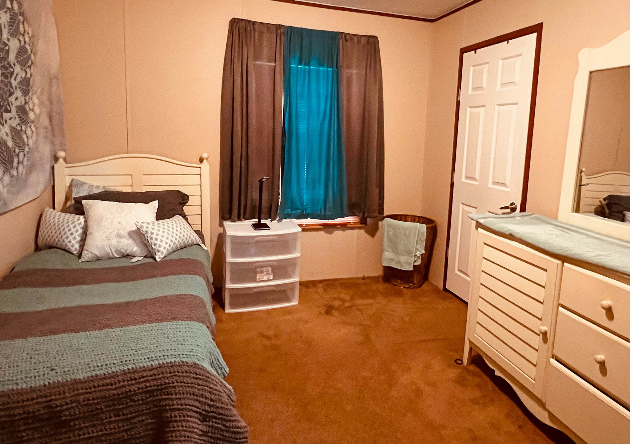 A small bedroom with a single bed, a dresser with a mirror, and a window between brown curtains inside the home of Group Home Providers Cory and Jessica Berger in Bedford County, Virginia.