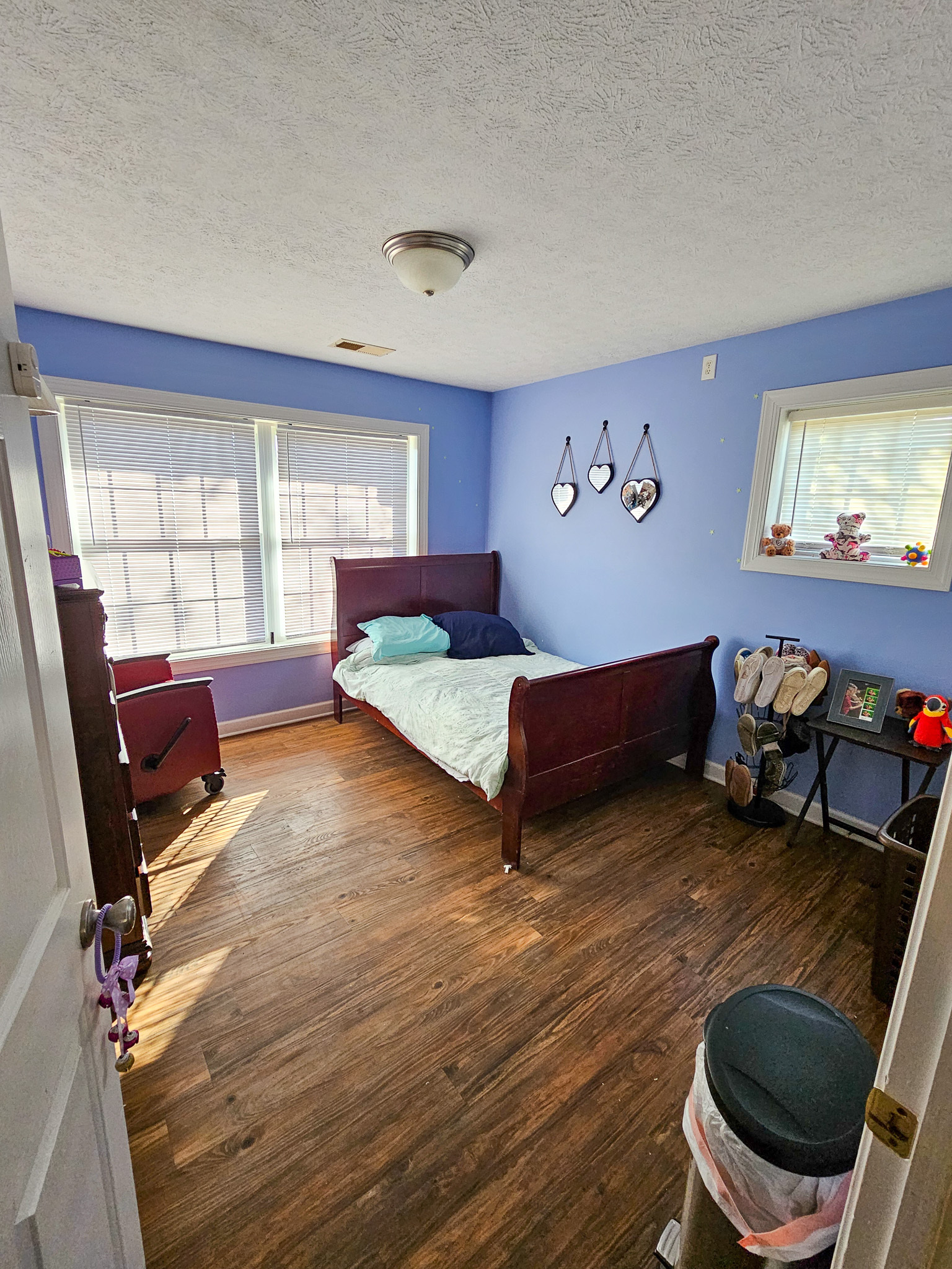 A bedroom with blue walls contains a wooden bed, a red armchair by the windows, and a small dresser with decorations at Boonsboro Group Home in Lynchburg, Virginia.