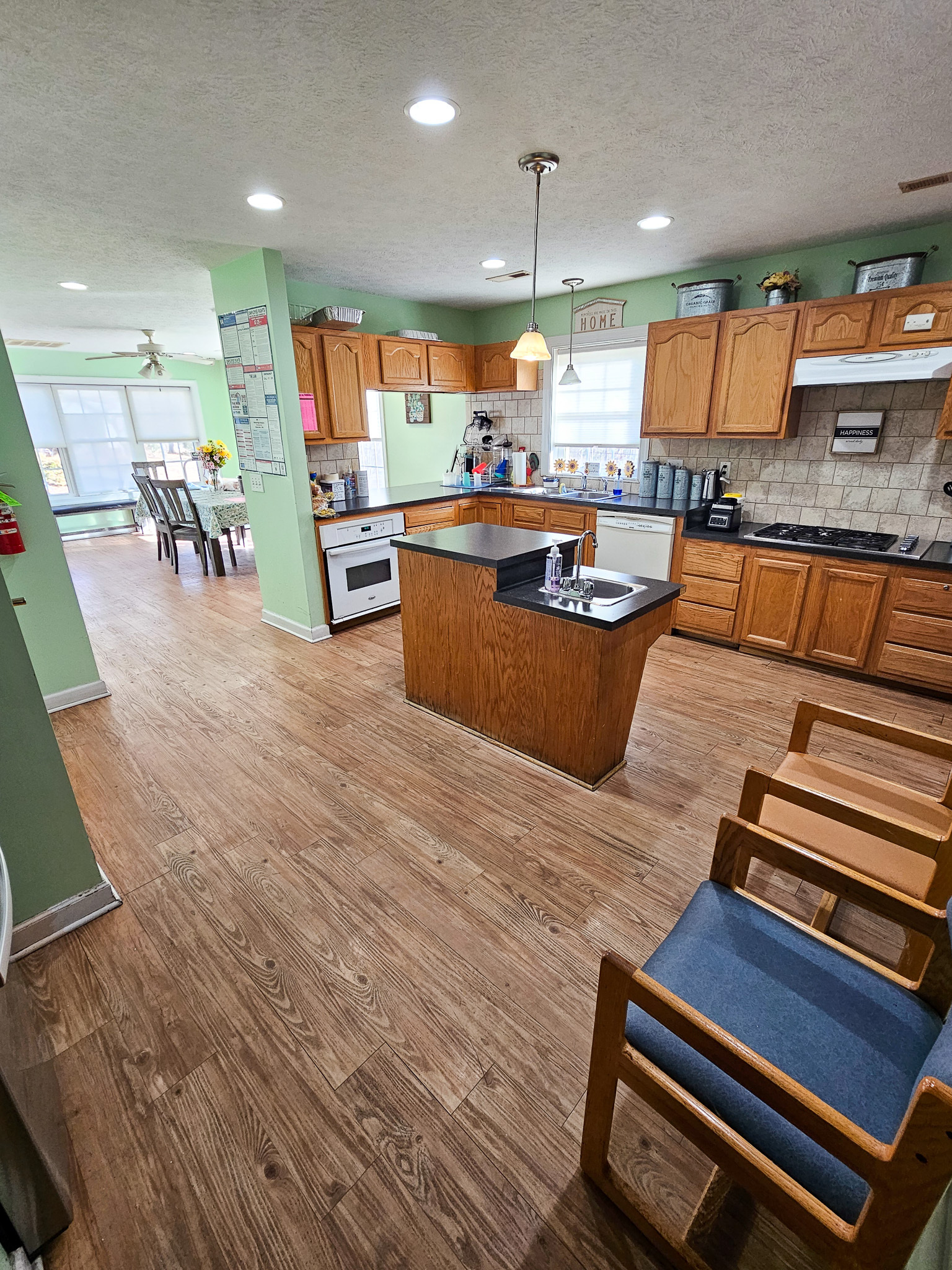 A spacious kitchen with light green walls features wooden cabinets, a center island, and a dining area visible in the background at Boonsboro Group Home in Lynchburg, Virginia.