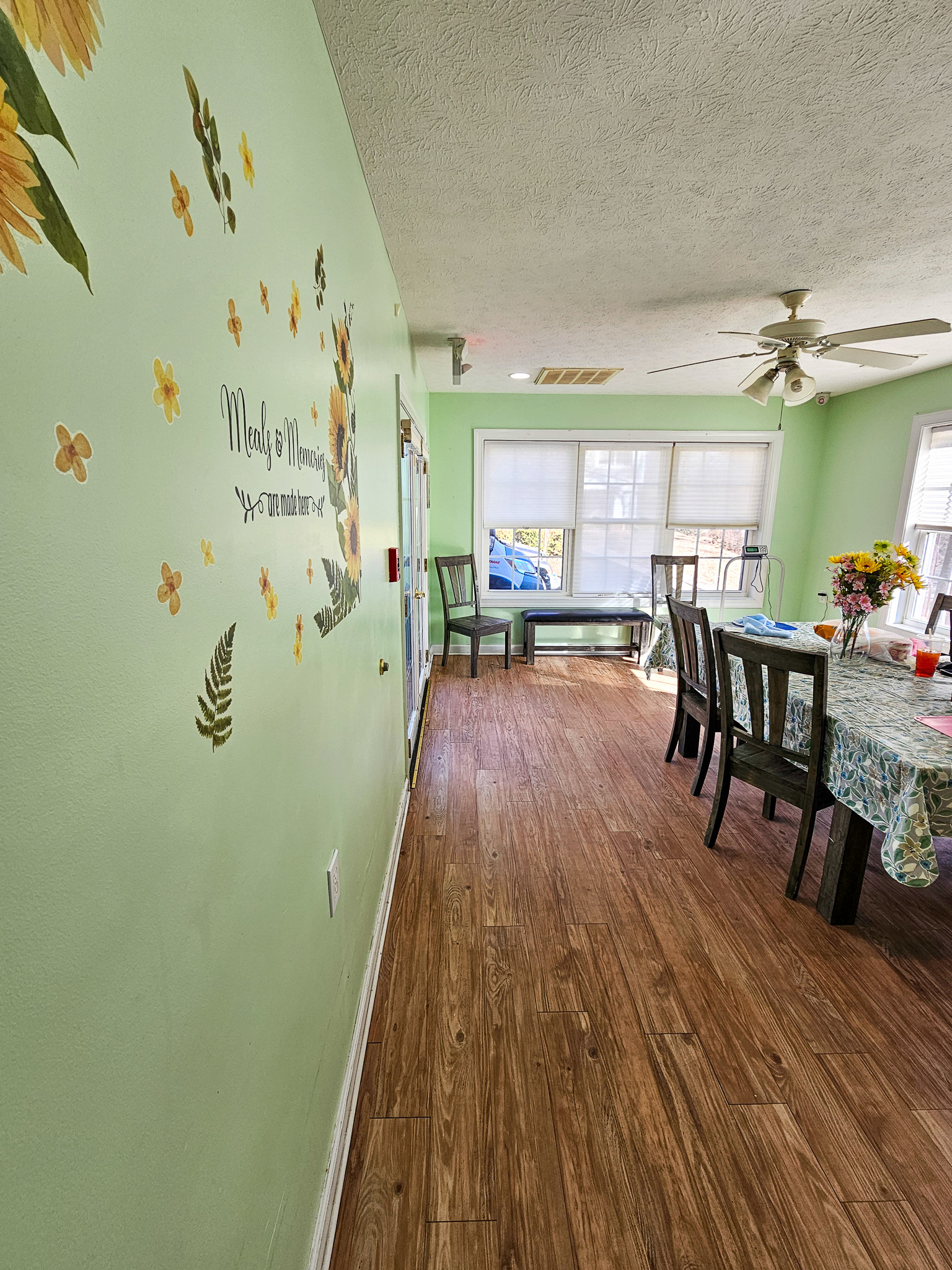 A dining area with light green walls features wall decals, a table with chairs, and large windows at the far end at Boonsboro Group Home in Lynchburg, Virginia.