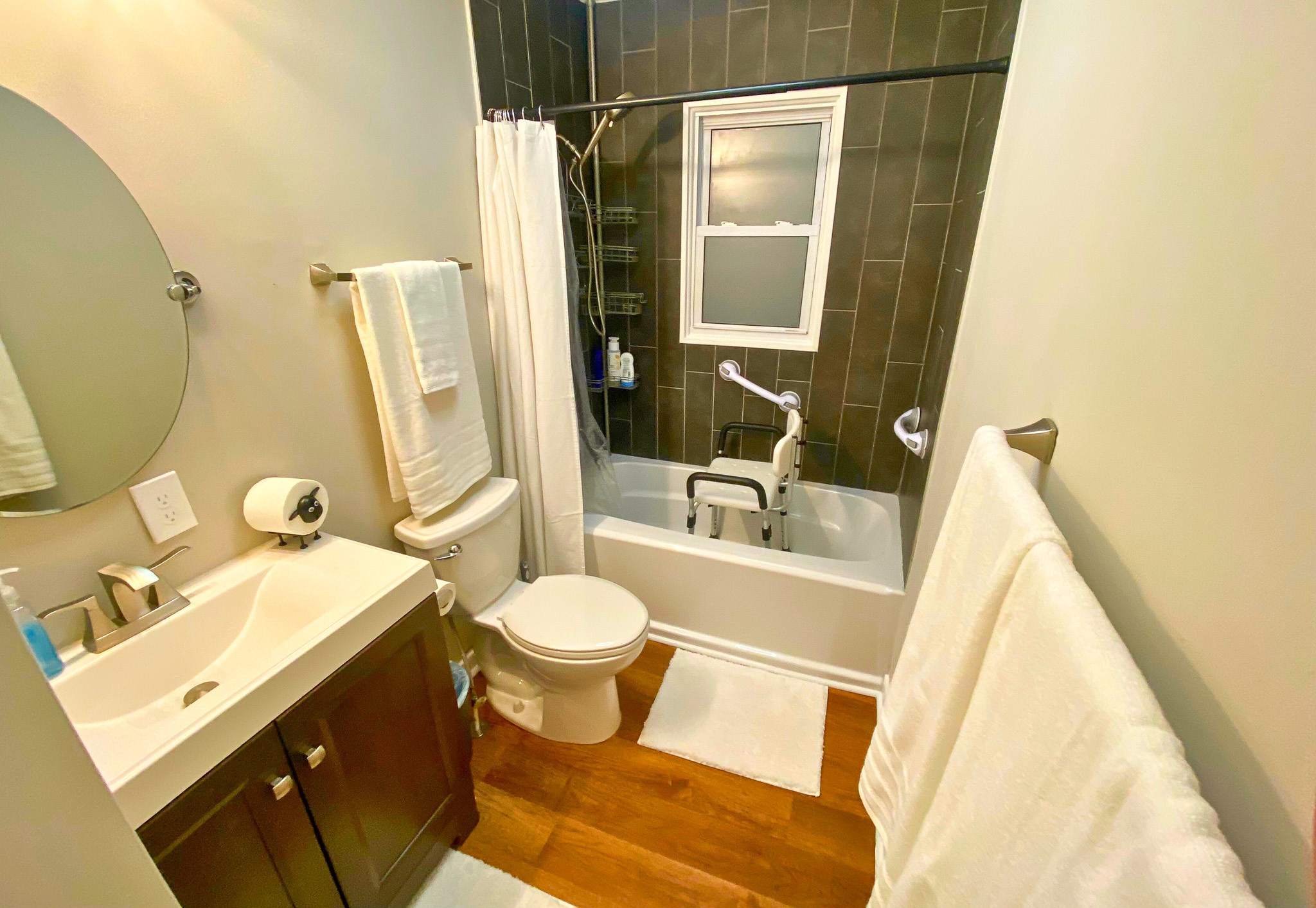 Bathroom with a vanity sink, toilet, and bathtub shower combination featuring dark tile walls, white curtain, small window, and neatly hung towels belonging to Sponsored Residential Providers Bradley and Andrea Smith in Richmond.