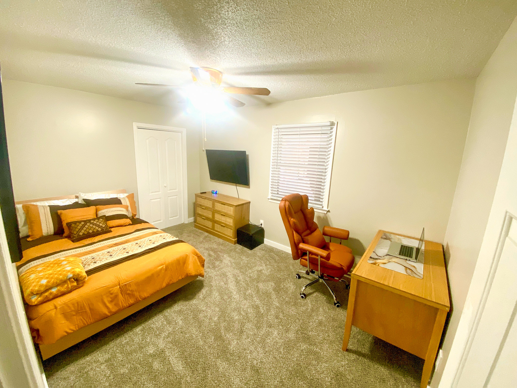 Bedroom with orange bedding on a wooden bed frame, dresser with TV, desk with chair, carpet flooring, and window with blinds beneath a ceiling fan and light belonging to Sponsored Residential Providers Bradley and Andrea Smith in Richmond.