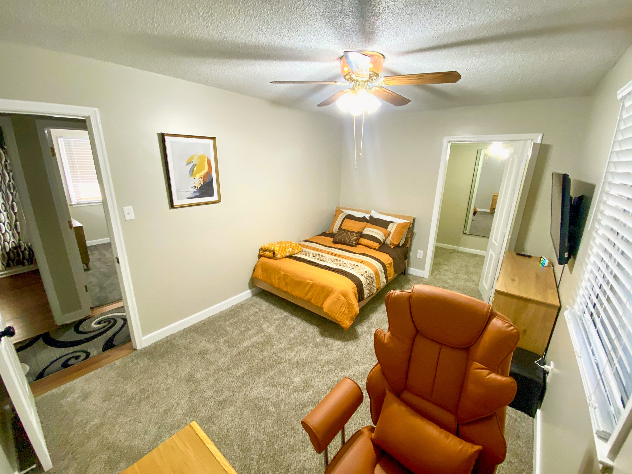 Bedroom with a bed featuring orange bedding, ceiling fan, dresser with TV, desk workspace, carpet flooring, and doorway to adjacent room belonging to Sponsored Residential Providers Bradley and Andrea Smith in Richmond.