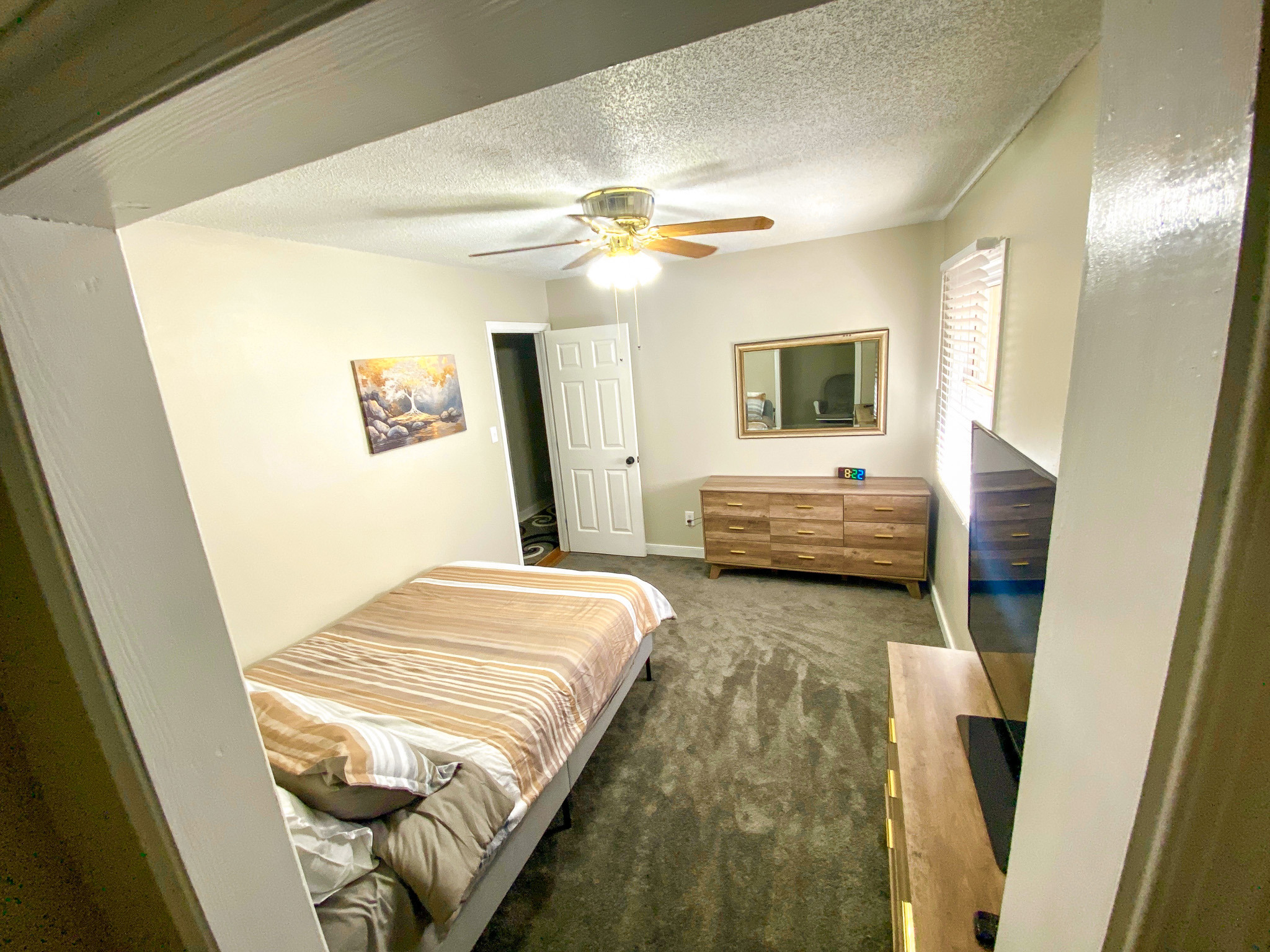 Bedroom with a wooden bed featuring striped bedding, dresser with mirror, wall-mounted TV, ceiling fan, carpet flooring, and doorway to hallway belonging to Sponsored Residential Providers Bradley and Andrea Smith in Richmond.