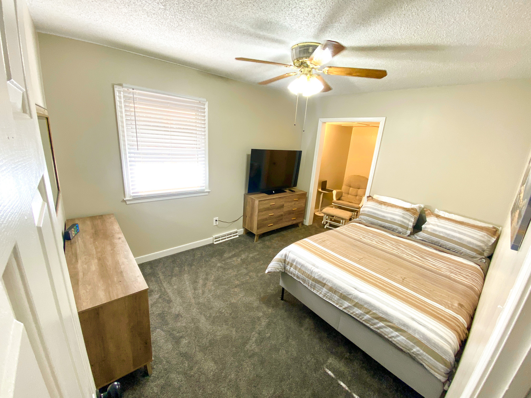 Light colored bedroom with striped bedding on a bed, wooden dresser, wall-mounted TV, ceiling fan, and window with blinds belonging to Sponsored Residential Providers Bradley and Andrea Smith in Richmond.