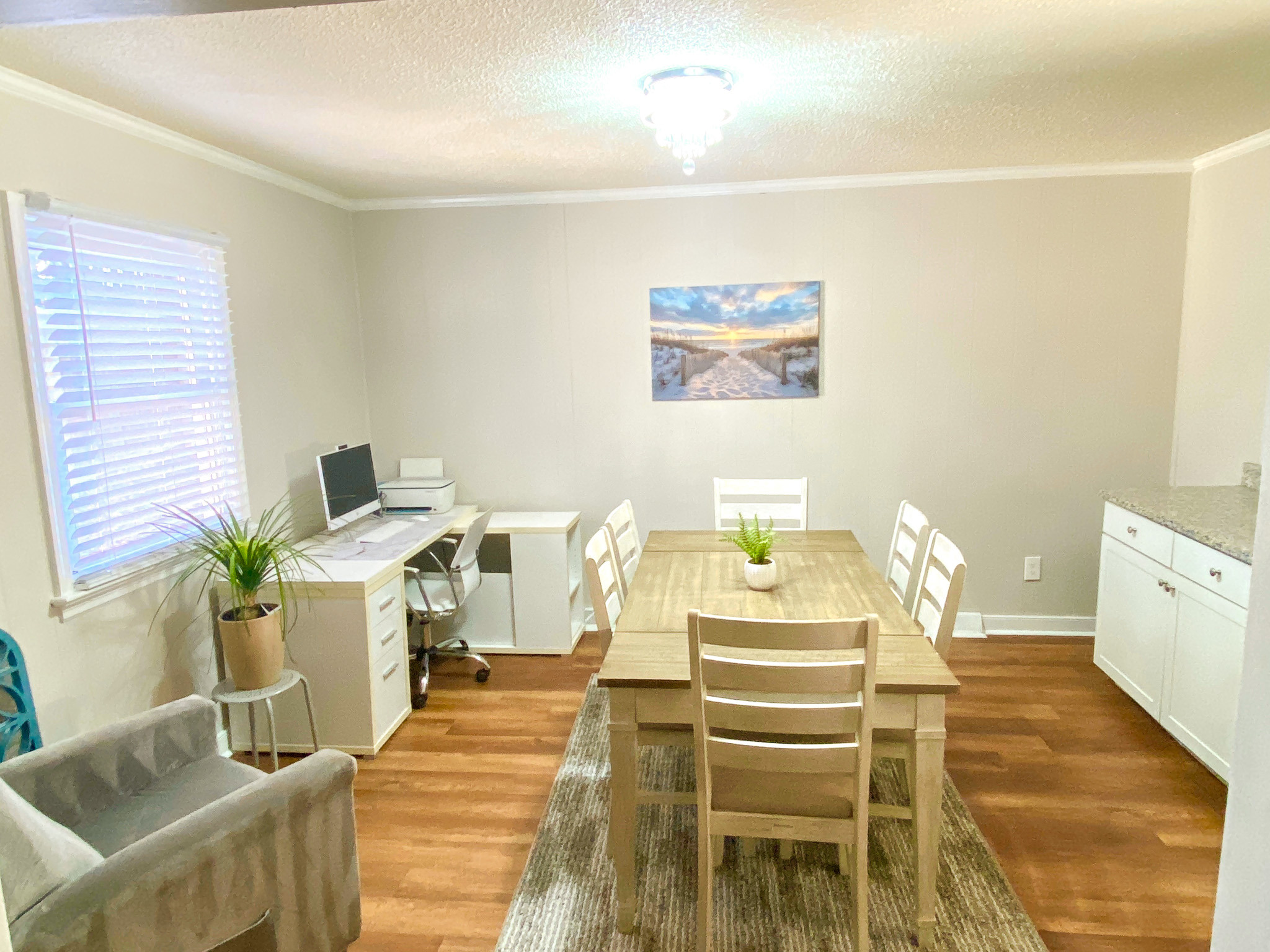 Dining room with a wooden table and six chairs on a patterned rug, white sideboard, small desk workspace, wall art, and light wood flooring beneath neutral walls belonging to Sponsored Residential Providers Bradley and Andrea Smith in Richmond.