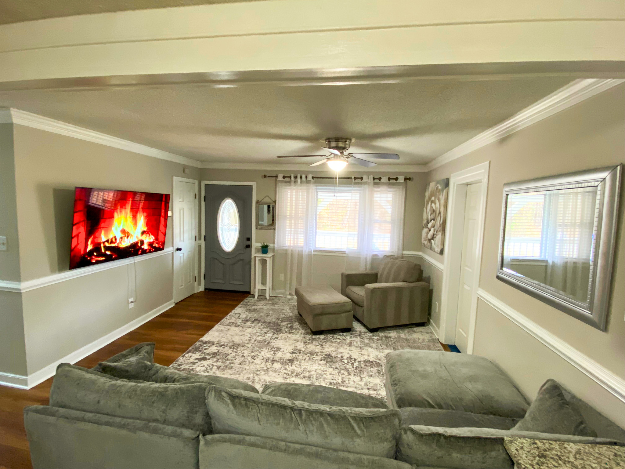 Open living room with gray sectional sofa, armchair, large area rug, wall-mounted TV displaying a fireplace, and hardwood floors beneath neutral walls belonging to Sponsored Residential Providers Bradley and Andrea Smith in Richmond.