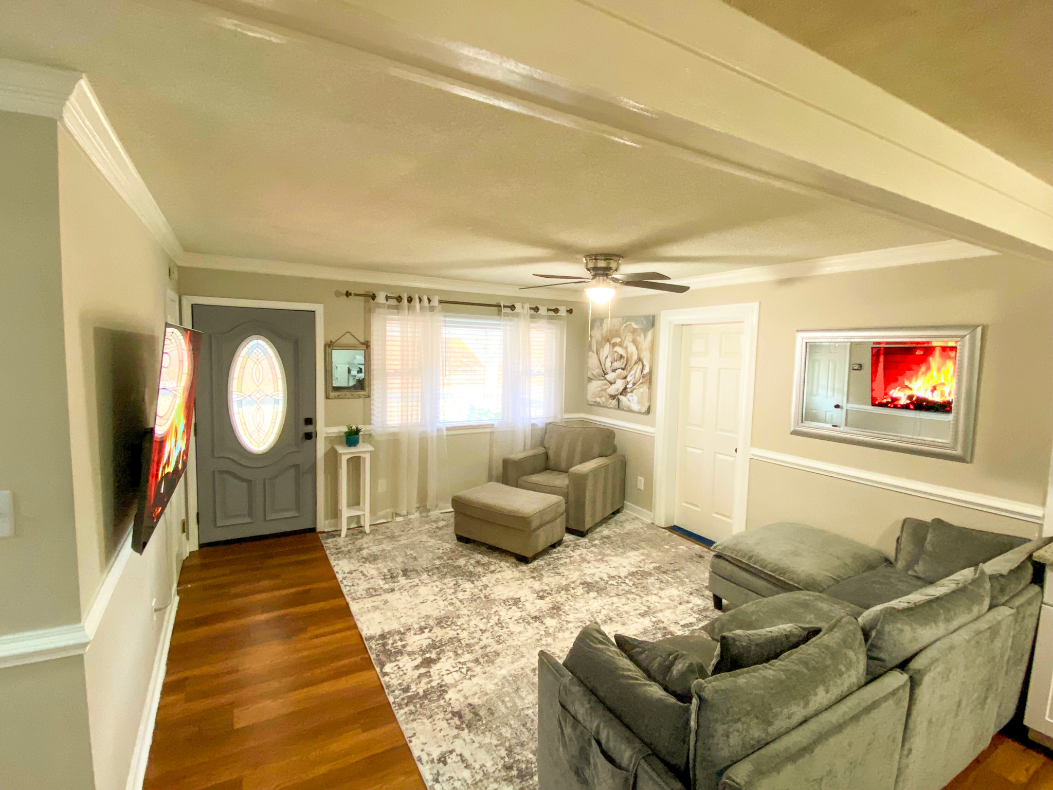 Open living room with gray sectional sofa, armchair, large area rug, hardwood floors, ceiling fan, front door with oval glass window, and wall-mounted TV displaying a fireplace belonging to Sponsored Residential Providers Bradley and Andrea Smith in Richmond.