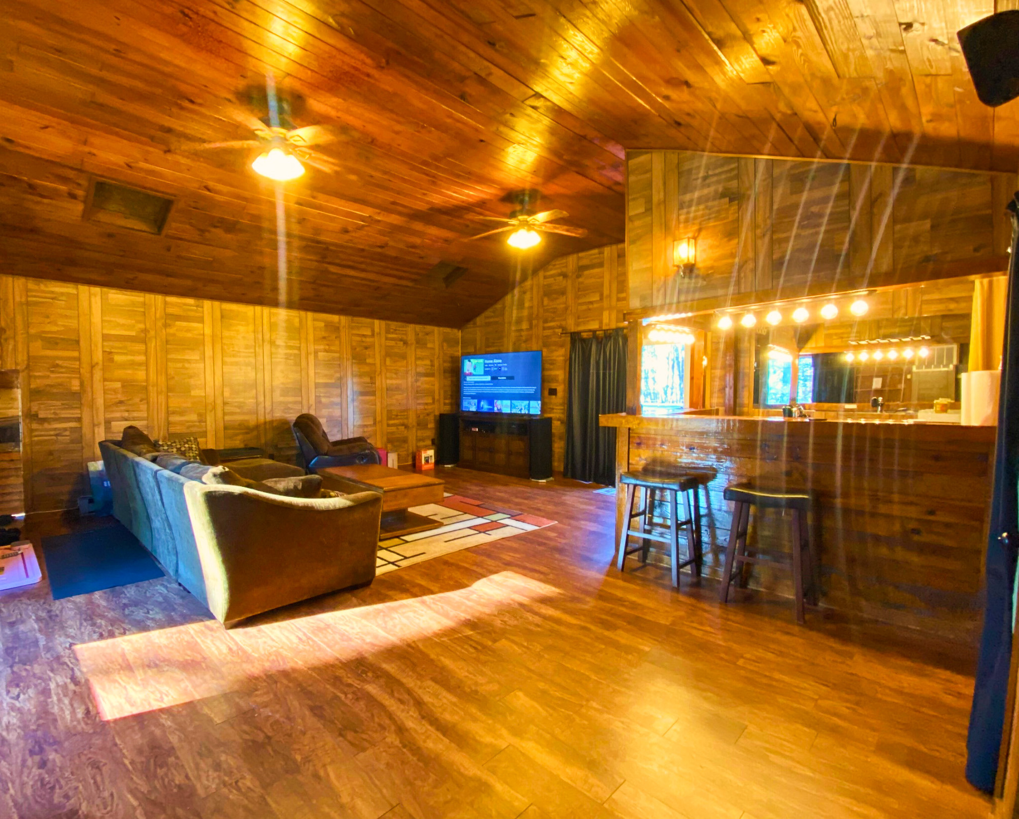 Wood-paneled upstairs rec room area with vaulted ceiling, sectional sofa, TV, bar counter with stools, and wood plank flooring illuminated by warm overhead lighting belonging to Sponsored Residential Providers Bradley and Andrea Smith in Richmond.