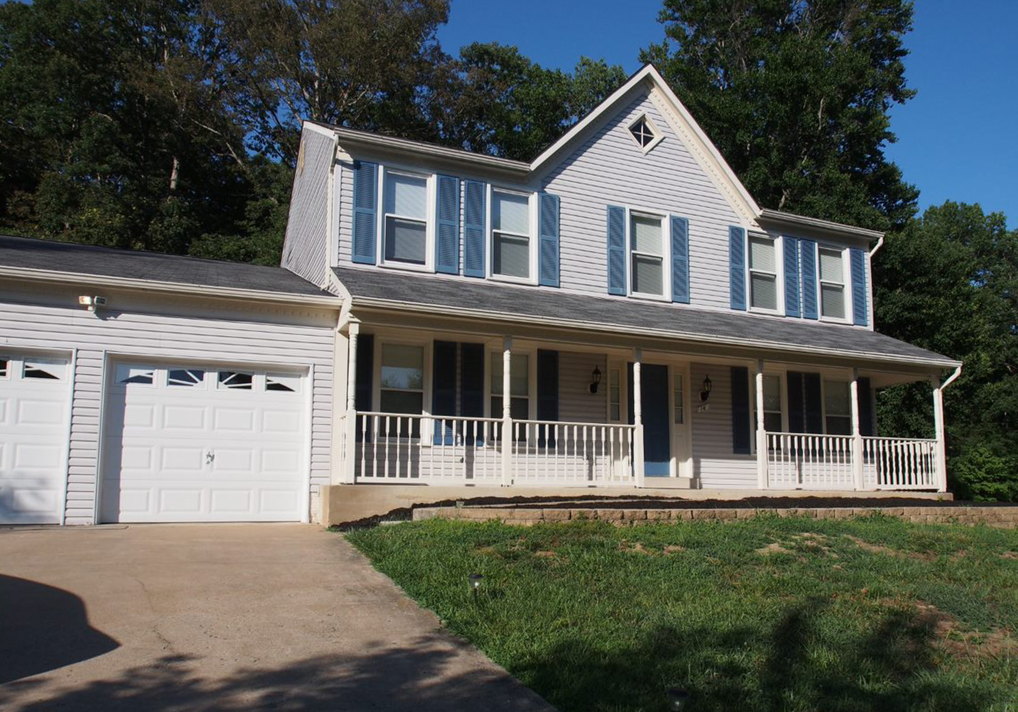 Two-story light-gray house with blue shutters, a front porch, and an attached two-car garage, set on a grassy lot with trees behind belonging to Sponsored Residential provider LaDonna Byrd in Northern Virginia.