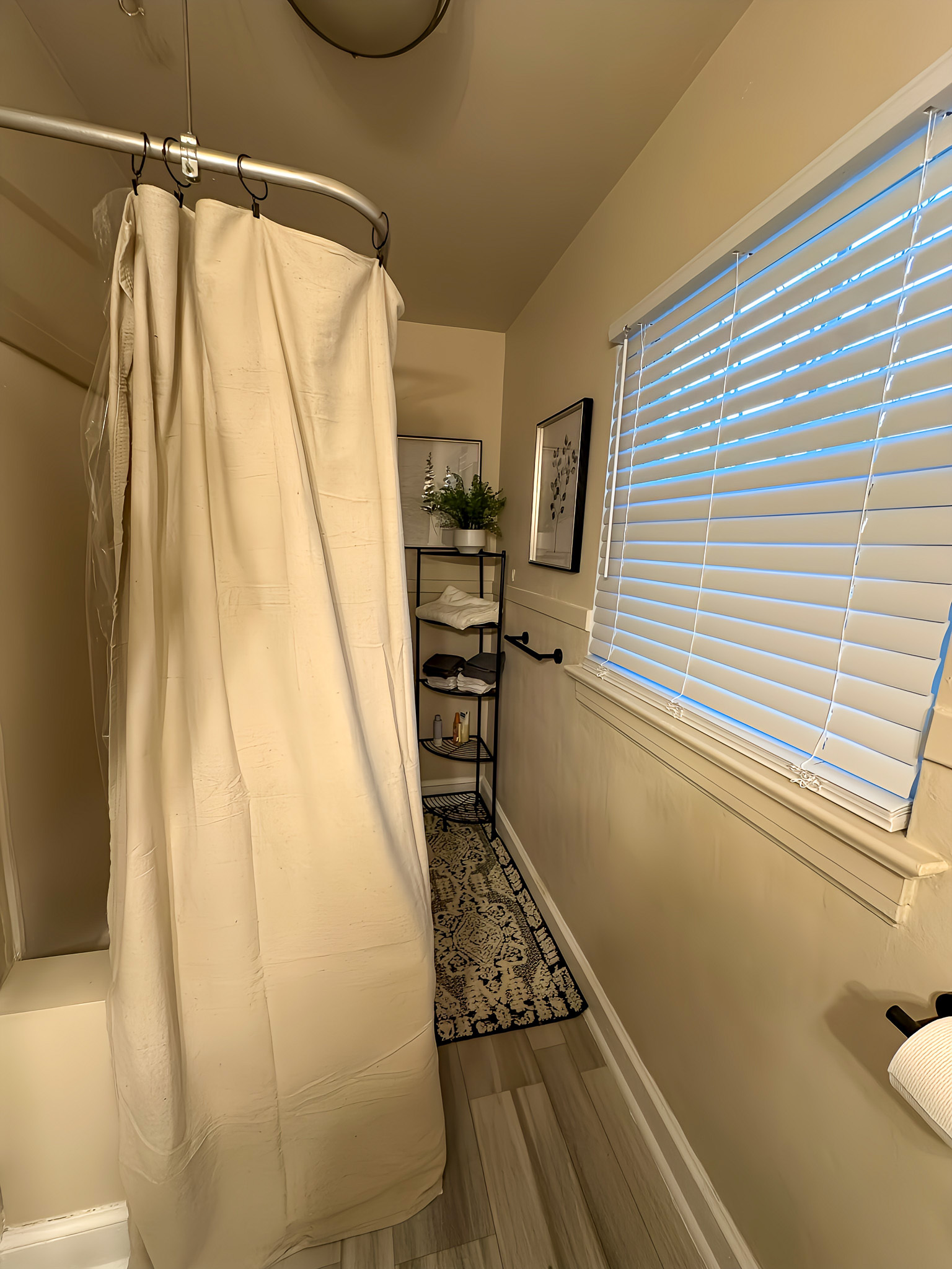 A narrow bathroom with a shower curtain on the left and a shelving unit holding towels and decor near the back wall inside the home of Sponsored Residential Providers Gia & Celia Calloway in Roanoke, Virginia.
