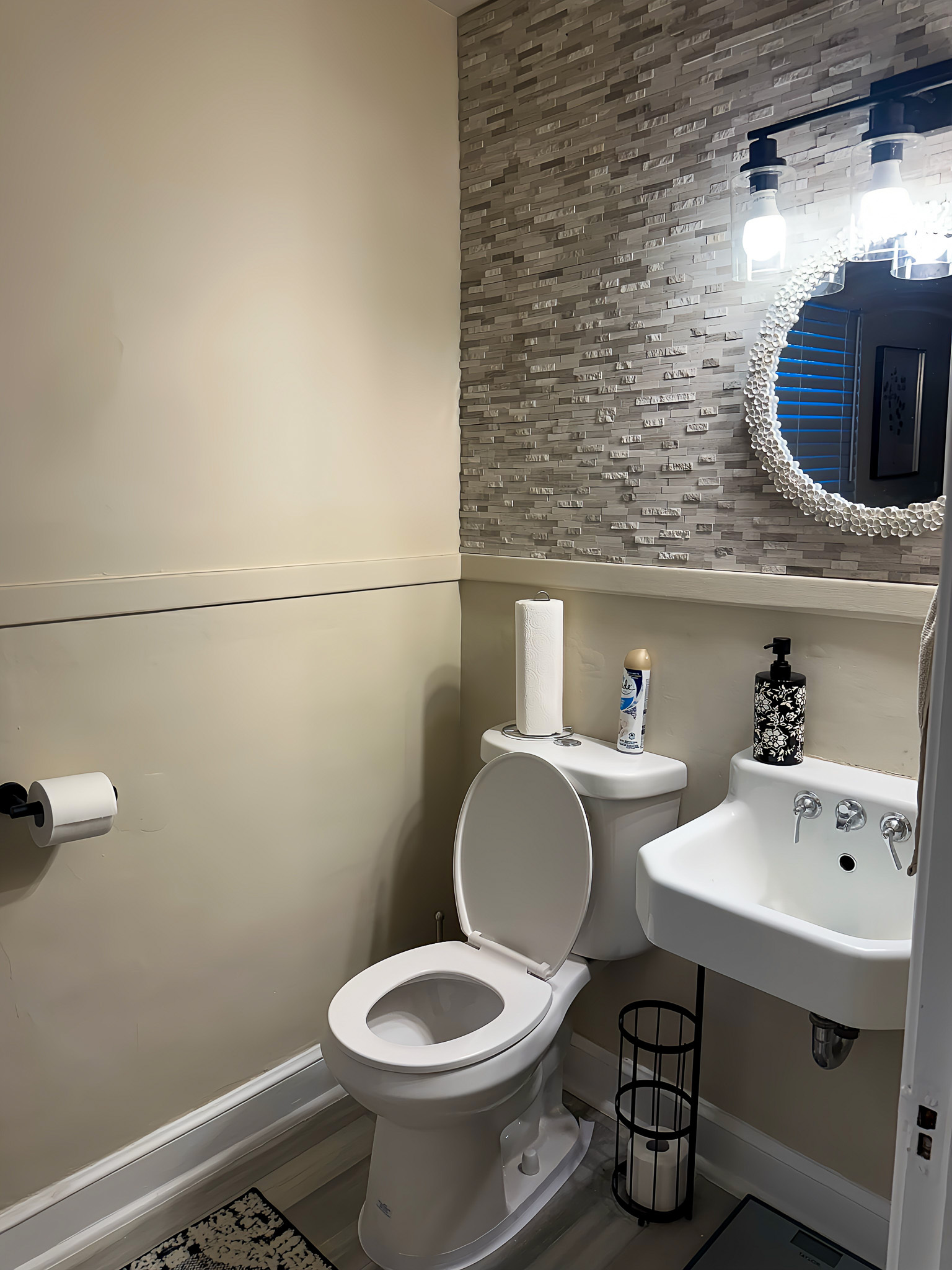 A small bathroom with a toilet, a wall-mounted sink, and a round mirror with lights above it inside the home of Sponsored Residential Providers Gia & Celia Calloway in Roanoke, Virginia.