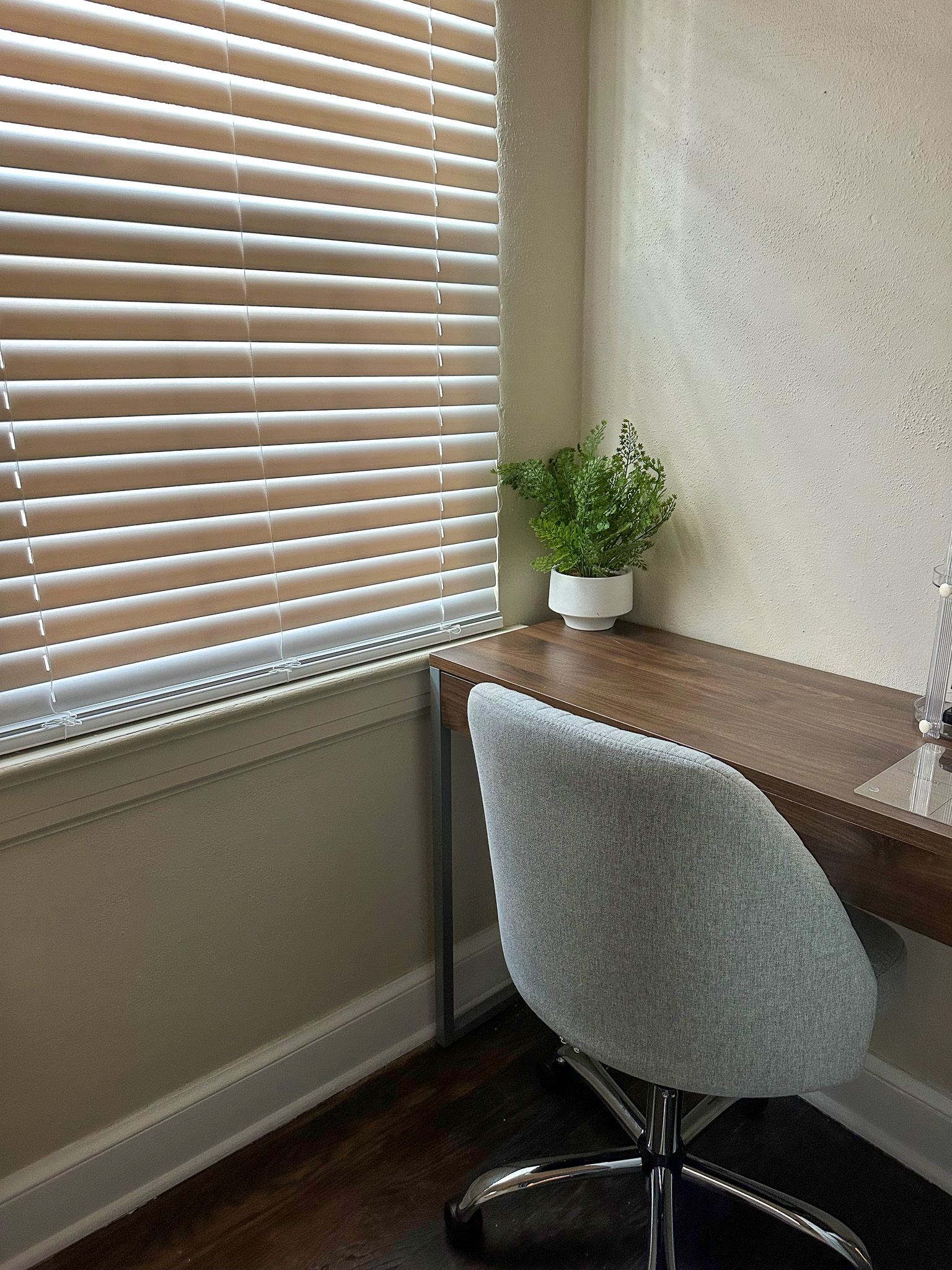A small workspace with a wooden desk, a gray swivel chair, and a potted plant beside a window with blinds inside the home of Sponsored Residential Providers Gia & Celia Calloway in Roanoke, Virginia.