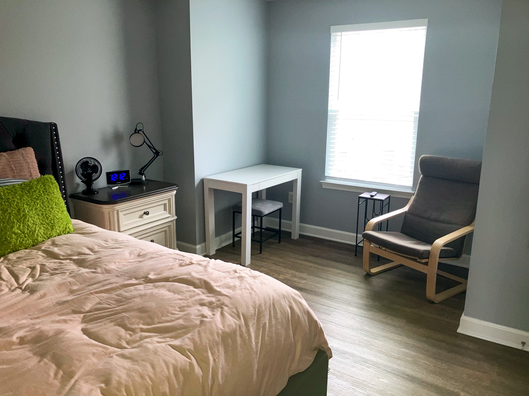 Bedroom with a bed, small nightstand, simple white desk with a chair, and an armchair near a window inside the home of Sponsored Residential Providers Crystal and Christopher Cameron in Fredericksburg, Virginia.