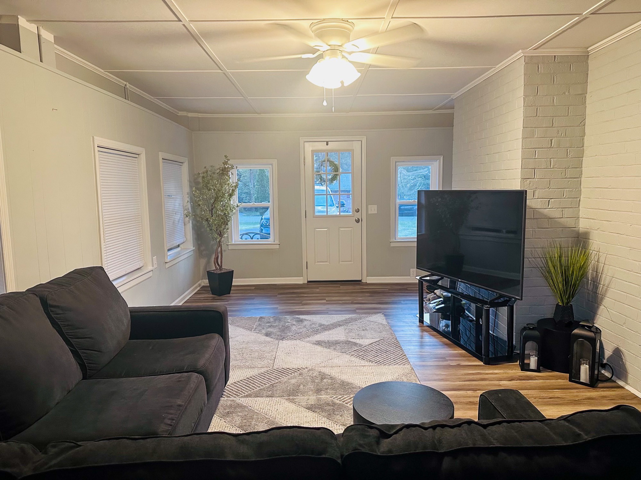 Spacious living room with sectional seating, TV on stand, brick accent wall, ceiling fan, and multiple windows and door allowing natural light inside the home of Sponsored Residential Providers Casey and Fred Doswell in Lynchburg, VA.