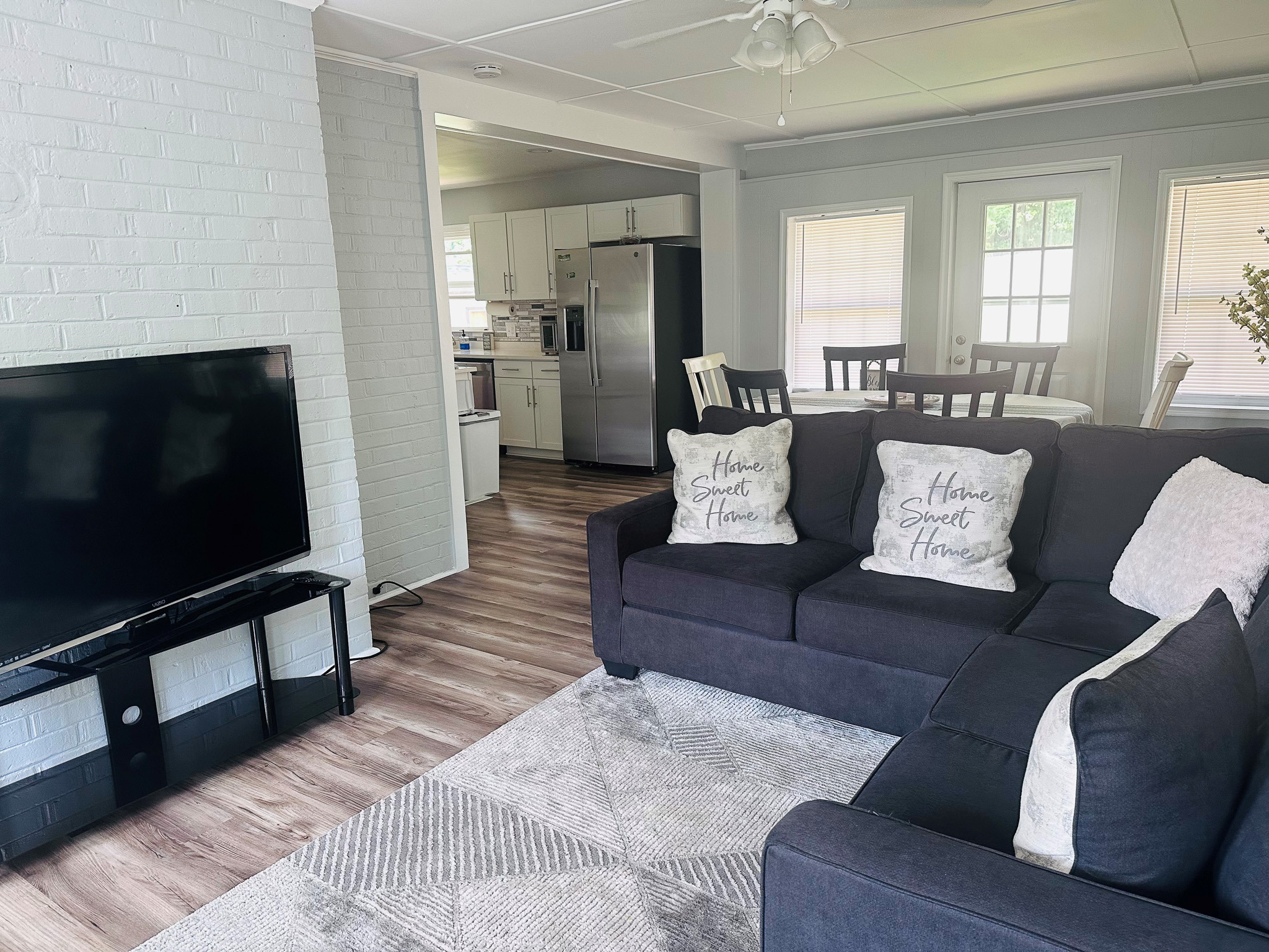Cozy living room with sectional sofa, TV, and rug, opening to a kitchen with white cabinetry and stainless steel refrigerator inside the home of Sponsored Residential Providers Casey and Fred Doswell in Lynchburg, VA.