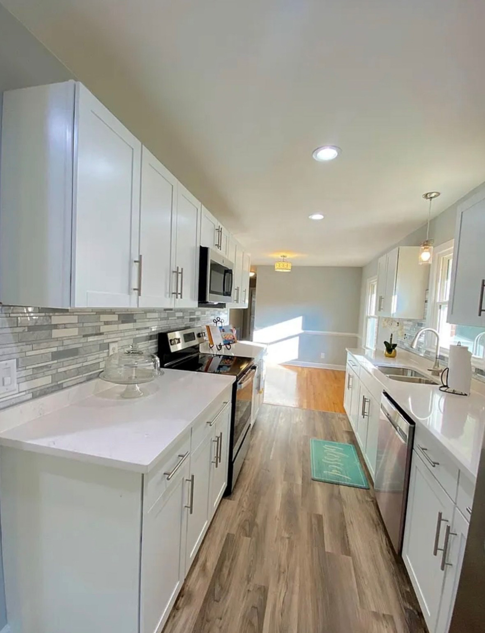 Bright galley-style kitchen with white cabinets, modern hardware, stainless steel appliances, tile backsplash, and wood-style flooring inside the home of Sponsored Residential Providers Casey and Fred Doswell in Lynchburg, VA.