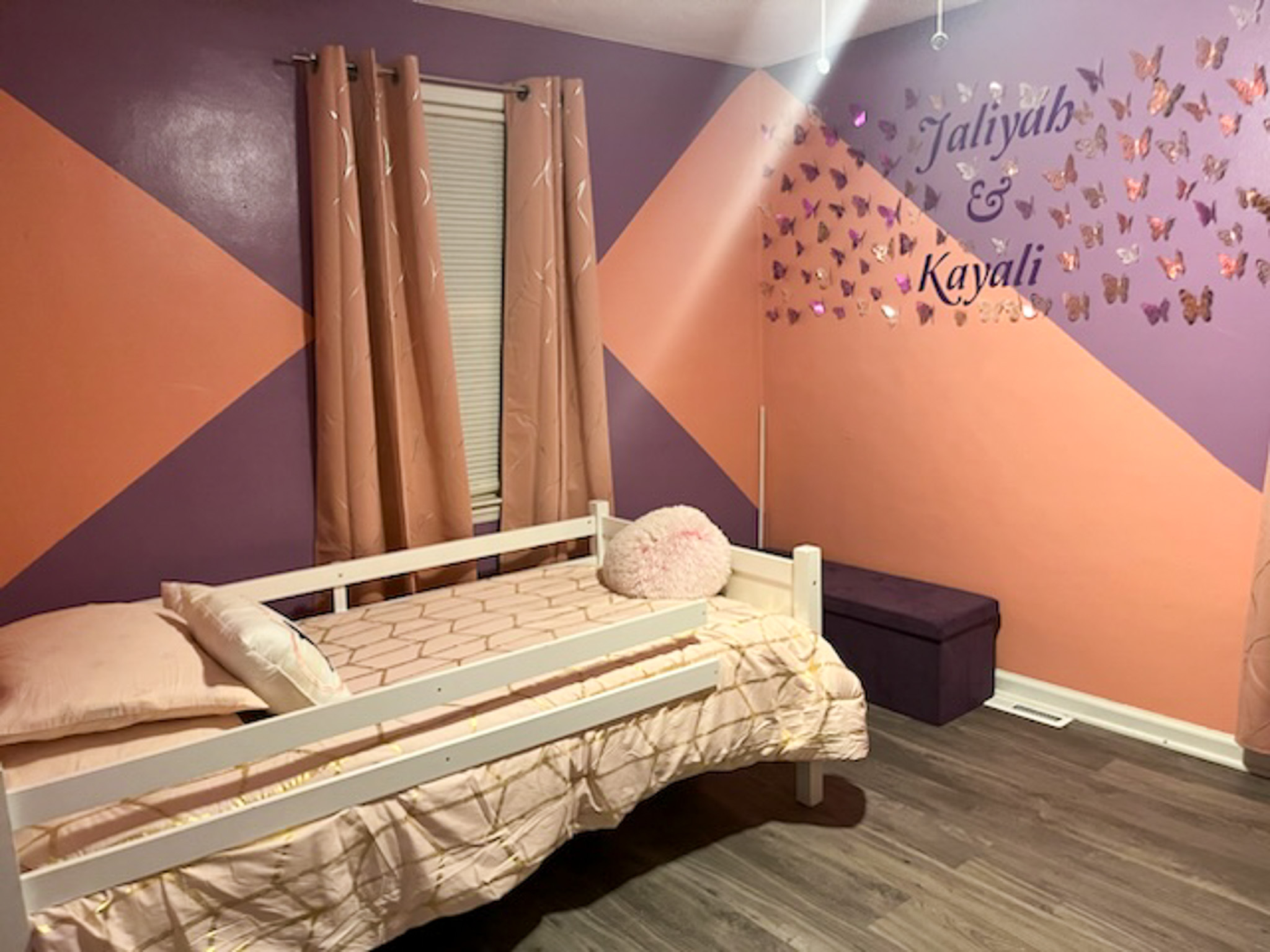 A colorful bedroom with pink and purple geometric walls, a small bed with a side rail, and decorative butterfly wall art above a storage bench inside the home of Sponsored Residential Provider Claudia Davis in Roanoke, Virginia.