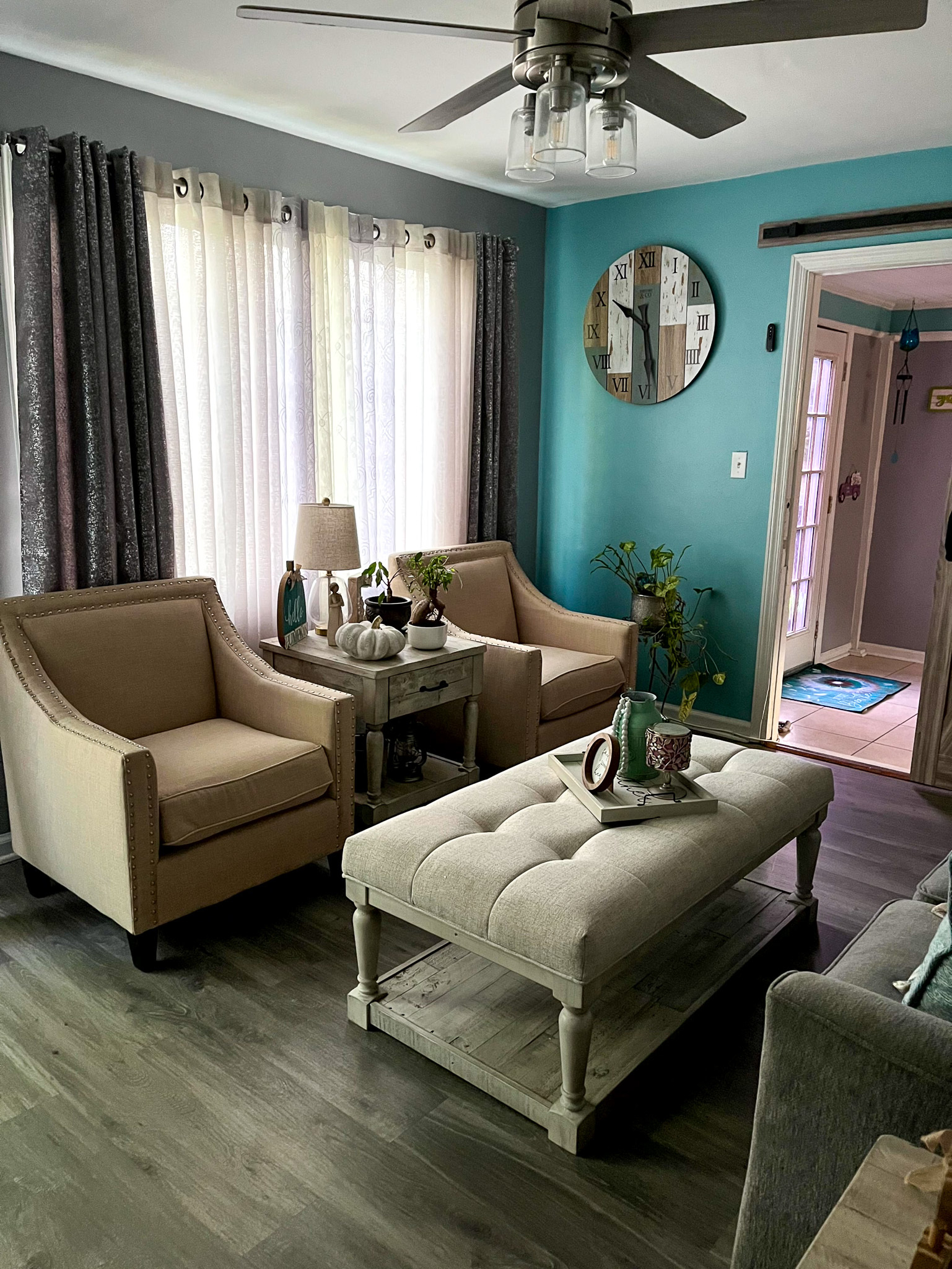 A sitting area with two beige armchairs, an upholstered coffee table, and a teal accent wall with a round mirror  inside the home of Sponsored Residential Provider Claudia Davis in Roanoke, Virginia.
