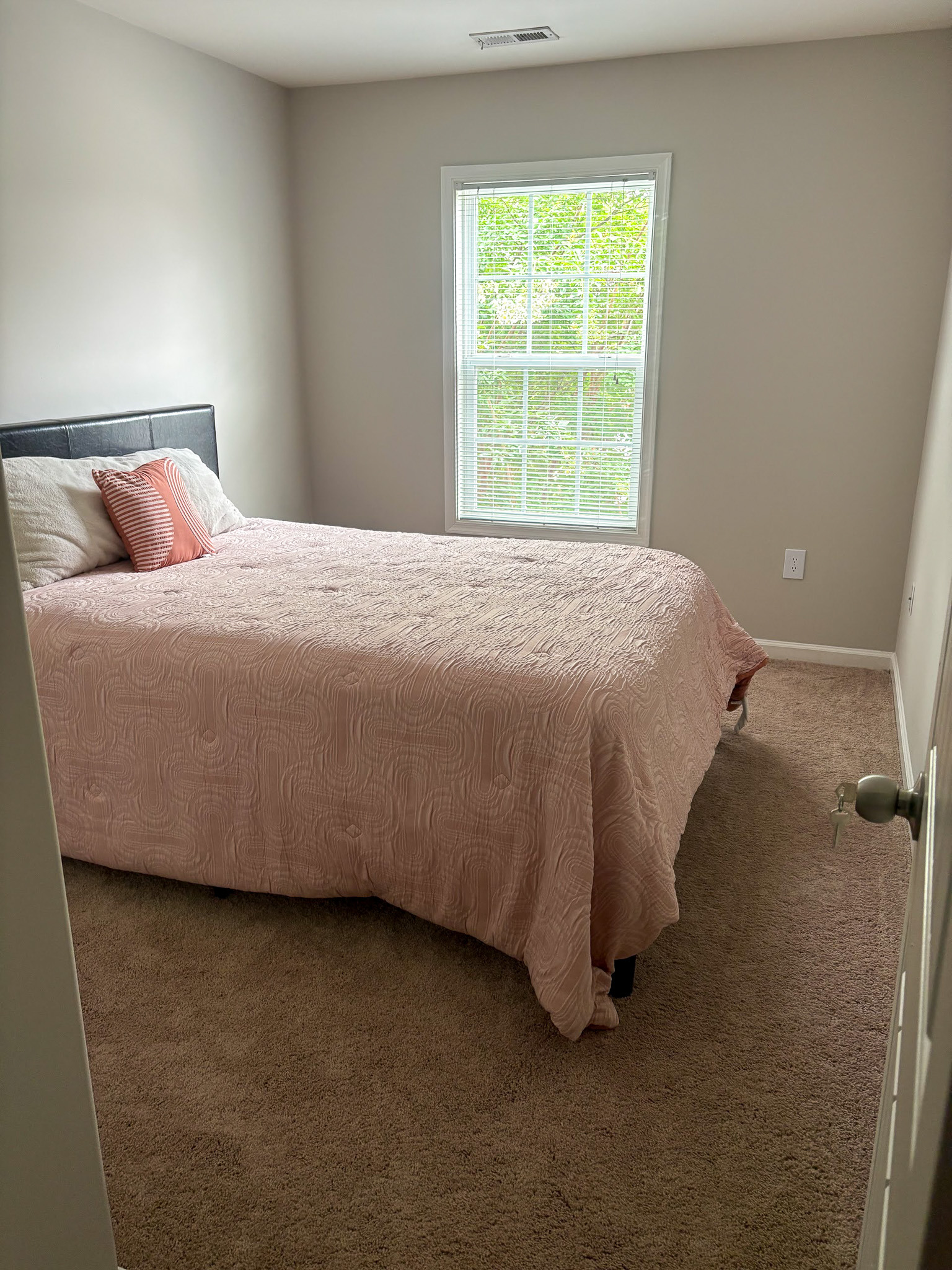 Bedroom with a queen-sized bed, neutral bedding, and a large window with blinds showing greenery outside inside the home of Sponsored Residential provider Cynthia Kimbrough-Mabry in Chester, Virginia