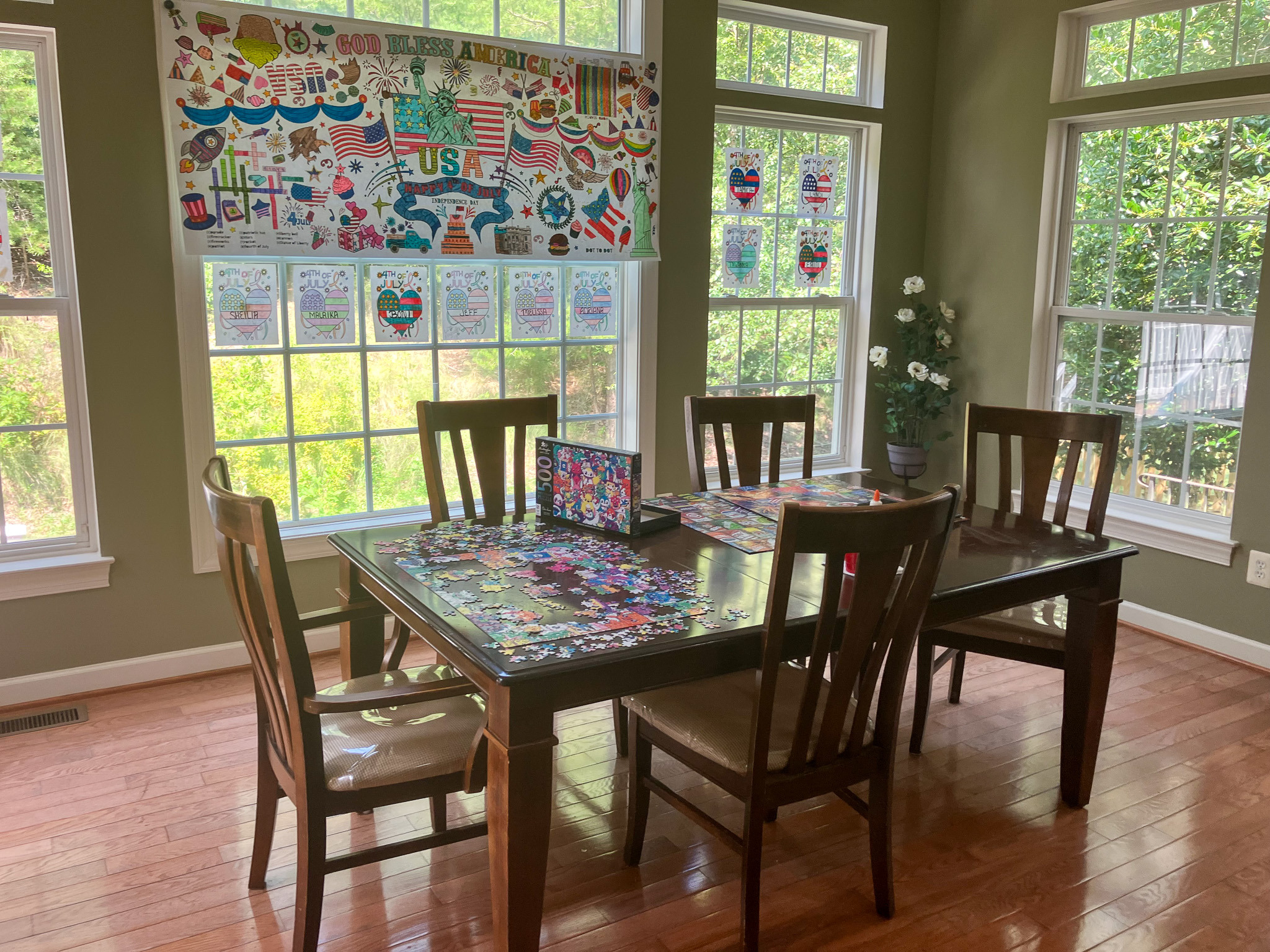 Dining room with wooden table, chairs, puzzle in progress, and large windows with colorful wall decorations belonging to the Agency operated group home named Deep Creek in Fredericksburg, Virginia.