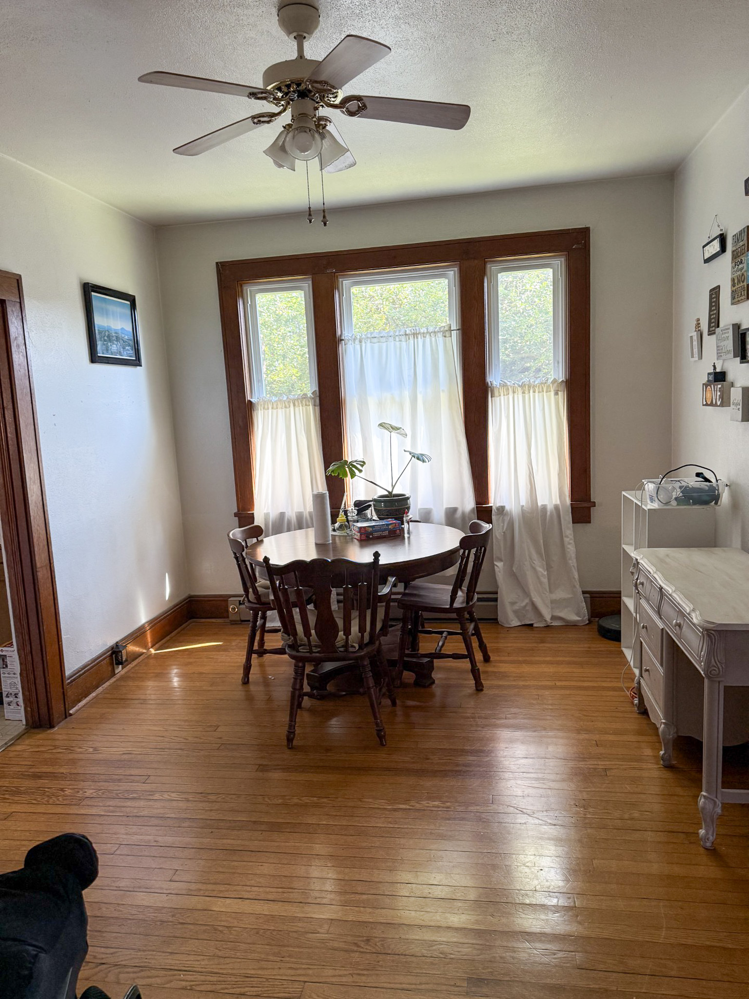 Bright dining room with wood floors, a round table and chairs, large windows with sheer curtains, and a ceiling fan overhead  inside the home of Sponsored Residential Provider Tammy Hunt in Floyd, Virginia.